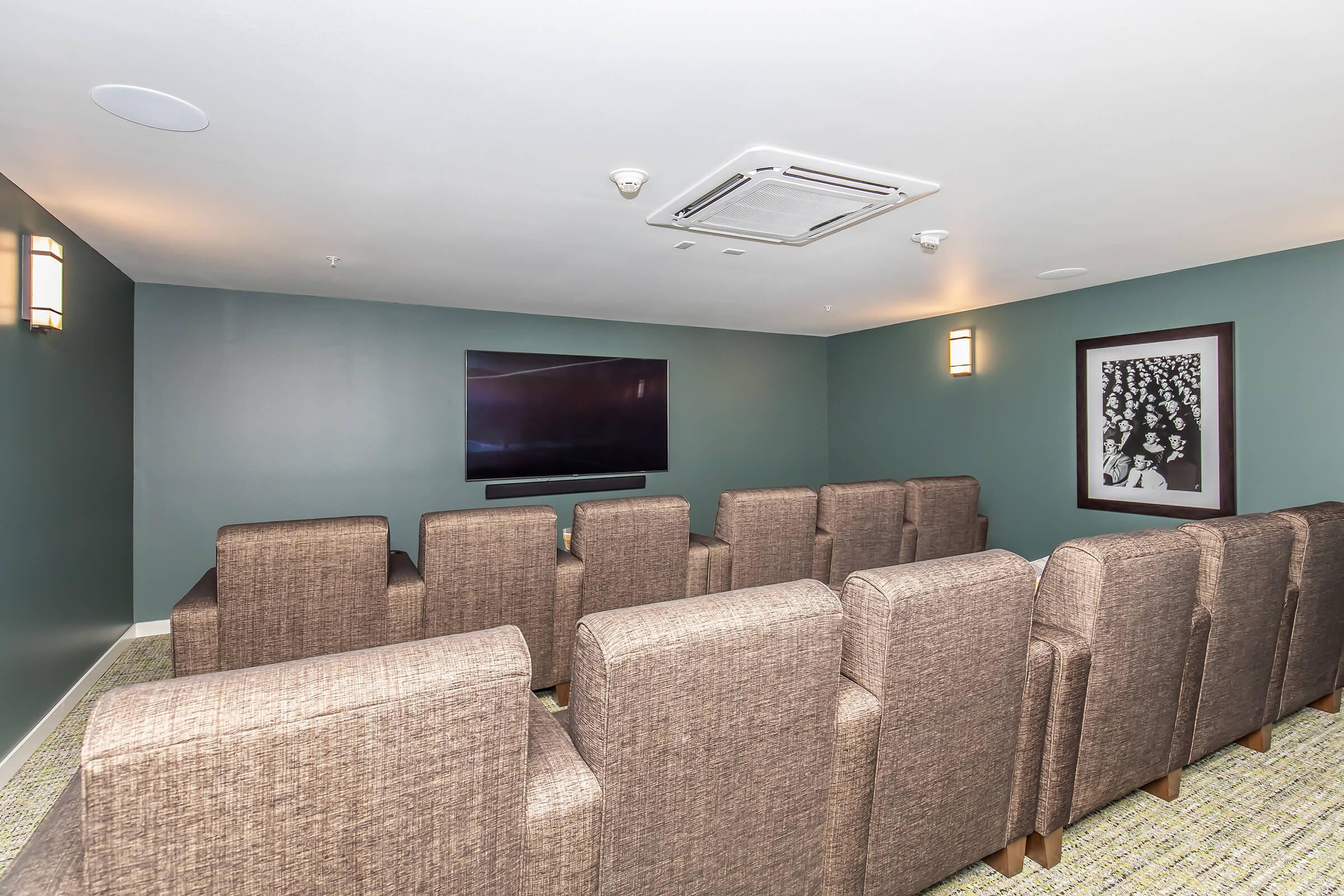 A small, modern media room featuring several upholstered chairs arranged facing a large flat-screen TV mounted on the wall. The walls are painted a teal color, and there’s a framed black-and-white photo on the adjacent wall. The space has ambient lighting and a neutral carpet.