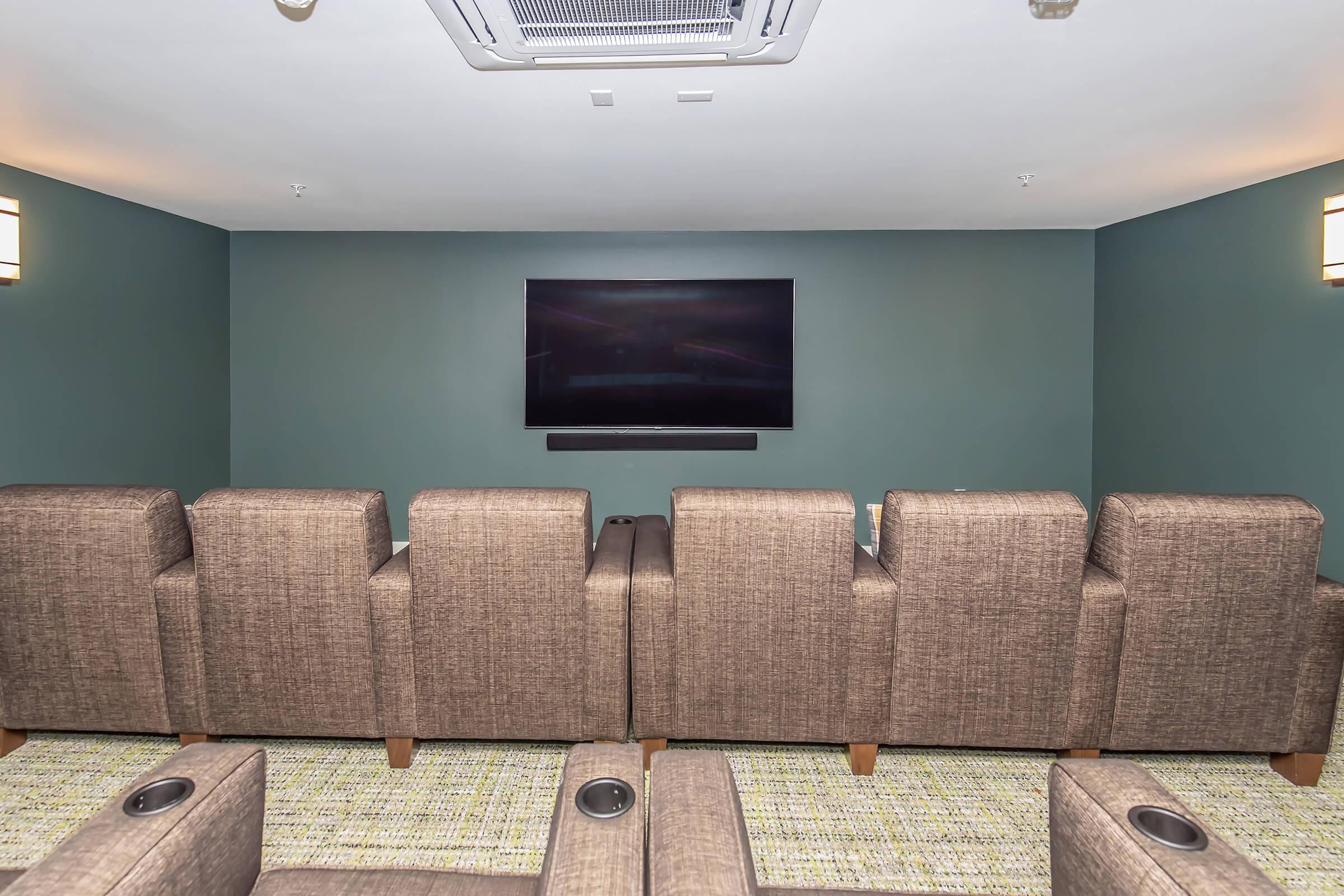 A cozy home theater setup featuring several plush, brown upholstered chairs arranged in front of a large flat-screen TV mounted on a dark green wall. The space is well-lit with wall sconces, and a textured carpet covers the floor.