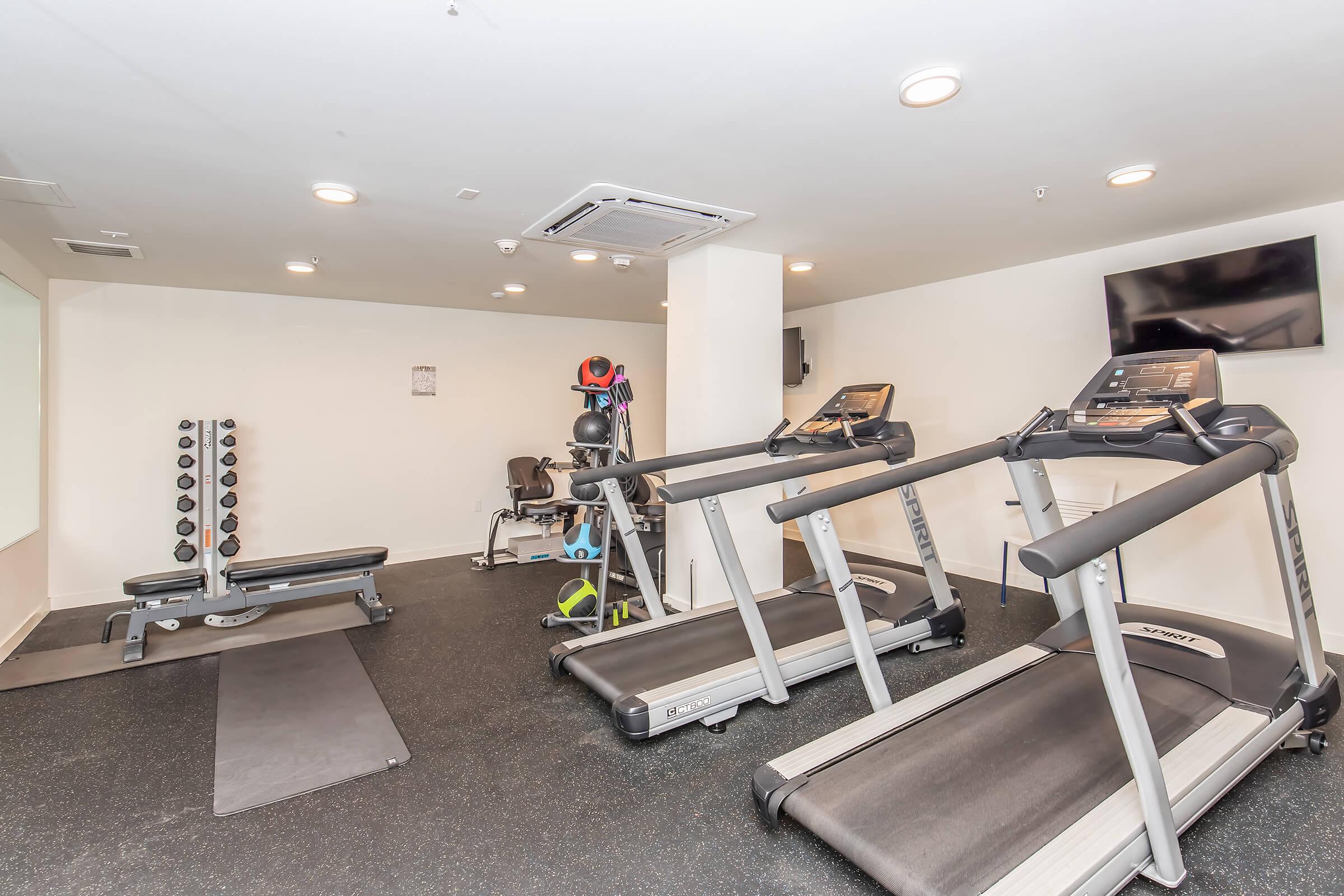 A modern gym interior featuring two treadmills, a weight bench with free weights, and various exercise accessories. The space has bright overhead lighting, a wall-mounted television, and a rubberized floor surface, creating an inviting environment for fitness activities.