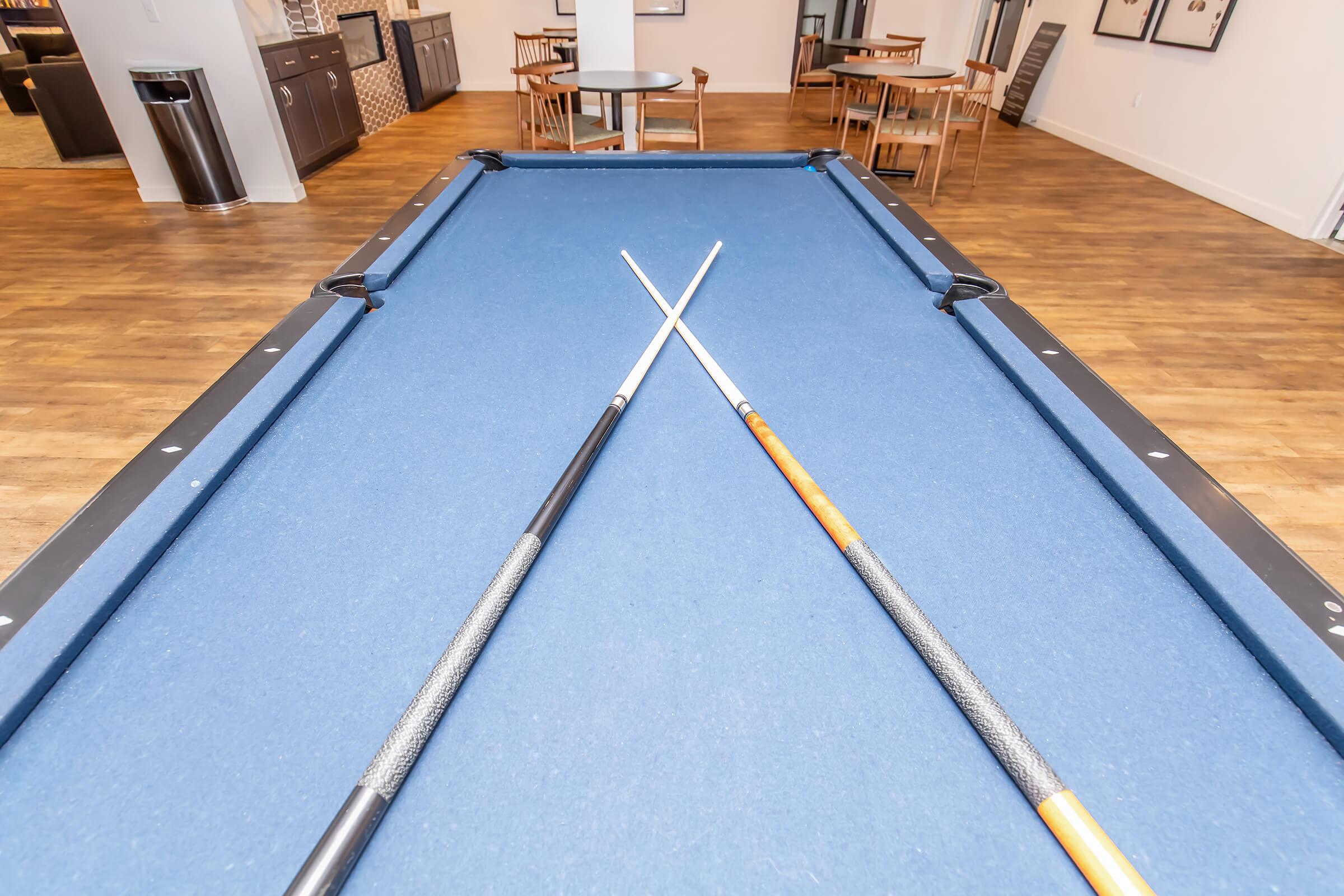 A blue billiard table with two crossed pool cues on top. The background features a modern recreational area with wooden flooring and tables.