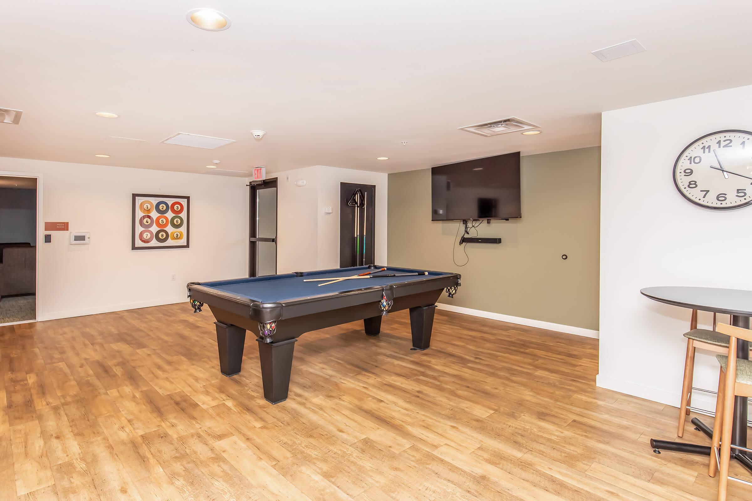 A modern game room featuring a pool table in the center, surrounded by wooden flooring. A large flat-screen TV is mounted on the wall, and a wall clock is visible. In the background, there are doors leading to another room, along with a framed artwork on the wall.