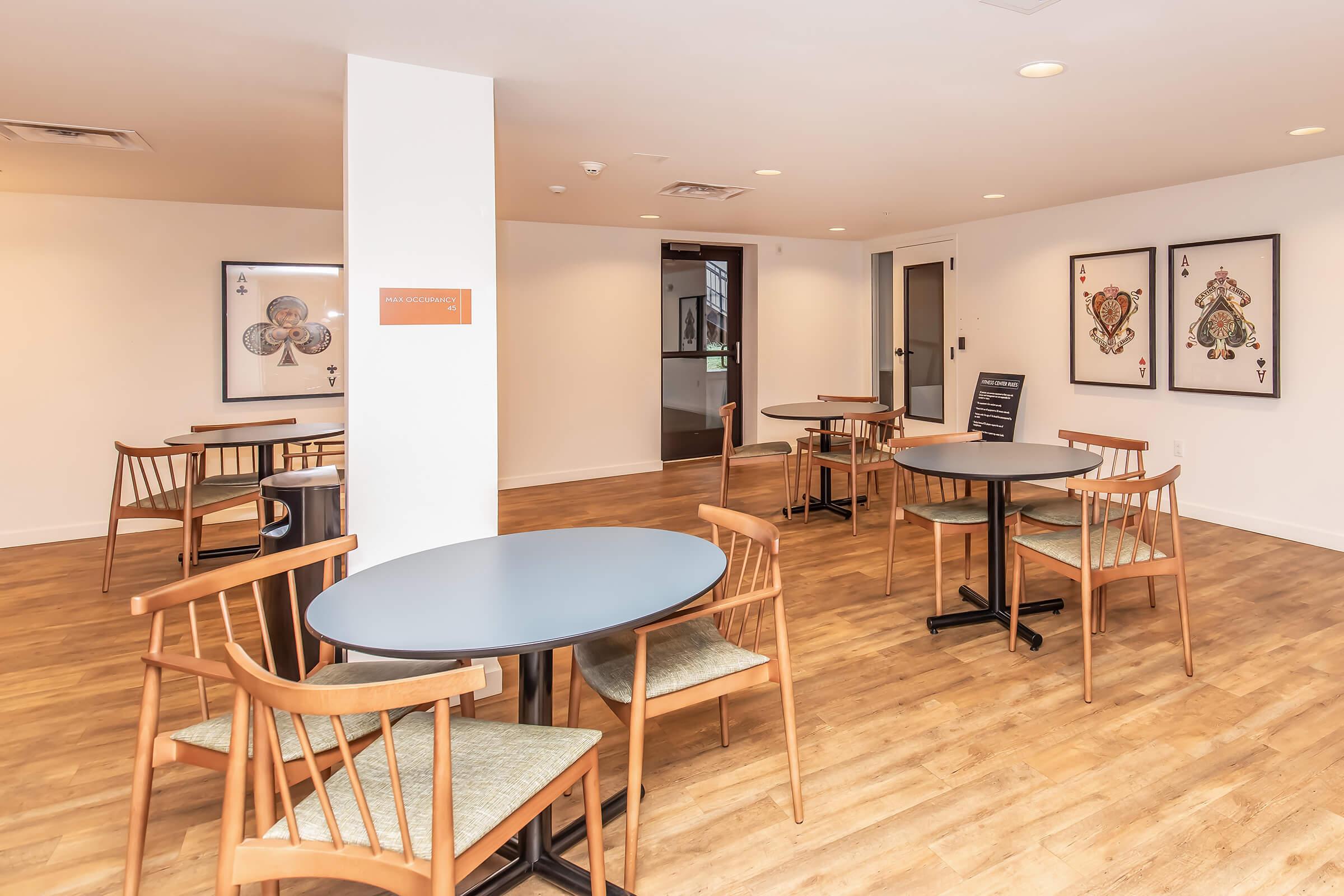A spacious common area featuring several round tables with wooden chairs arranged on a light wooden floor. The walls have framed artwork, and there is a sign on one wall. The room is well-lit with natural light coming through windows.