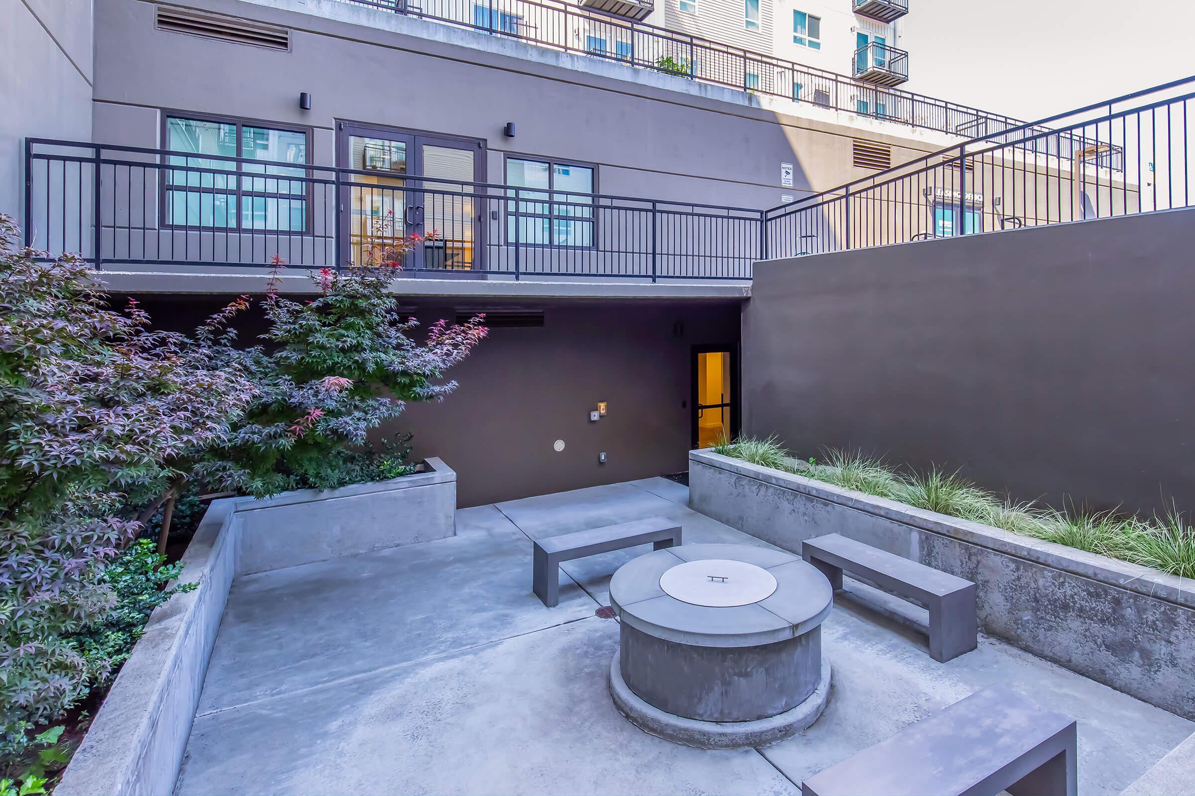 A modern outdoor courtyard featuring a circular concrete fire pit, surrounded by square seating benches. Lush greenery and shrubs are present along the edges, with a building facade in the background and balcony access above. The area appears stylish and designed for relaxation or gatherings.