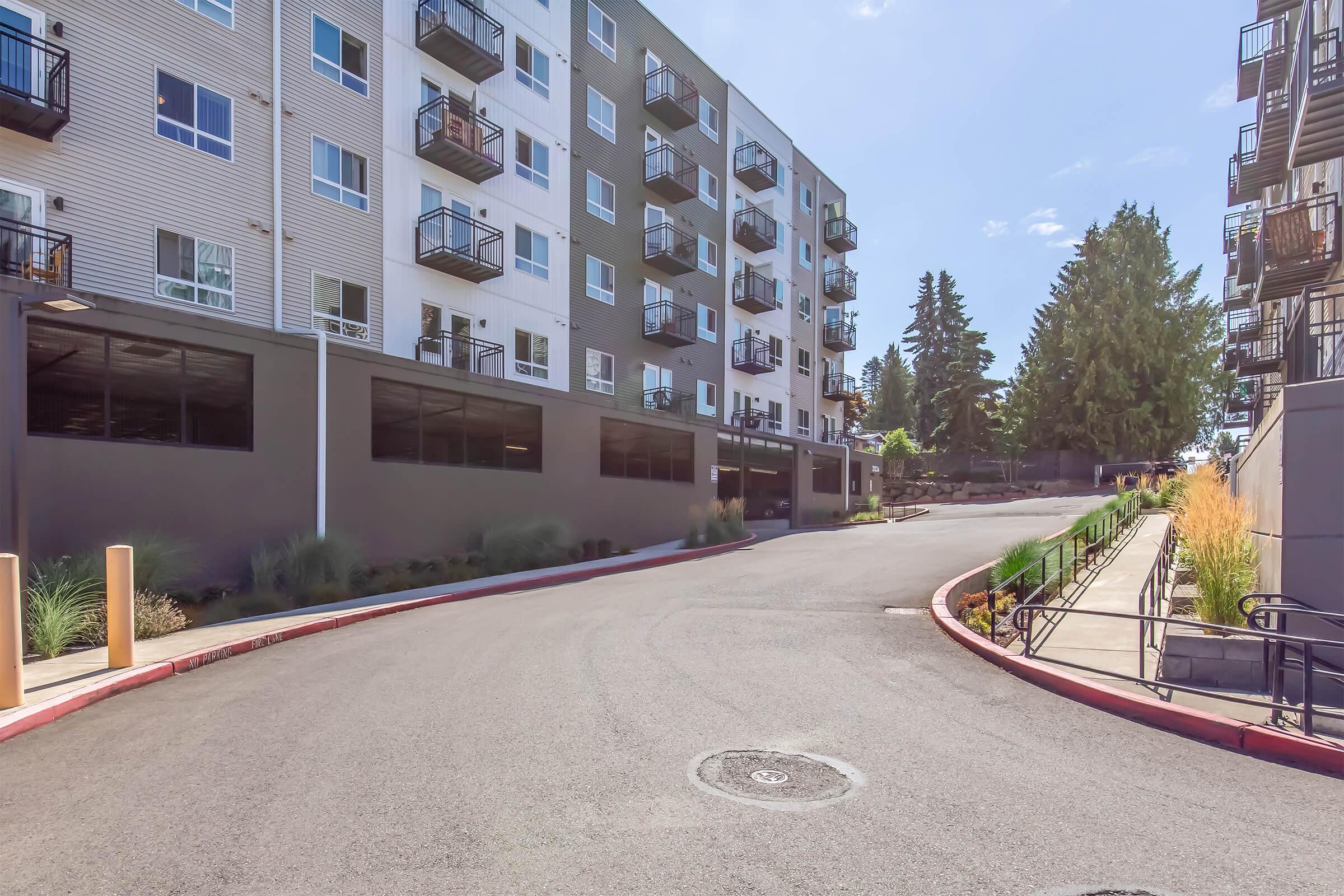 A view of a residential building with multiple stories, featuring balconies. The image shows a curved access road leading up to the entrance, flanked by landscaped greenery. Clear skies and trees in the background indicate a sunny day.