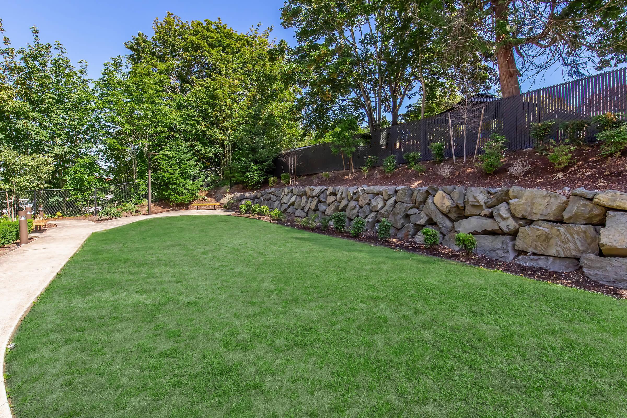 A well-maintained green lawn surrounded by trees and a stone wall. The area features a winding pathway and is designed for recreation, with seating and landscaping that enhances the natural beauty. Bright blue sky visible above, creating a serene outdoor space.