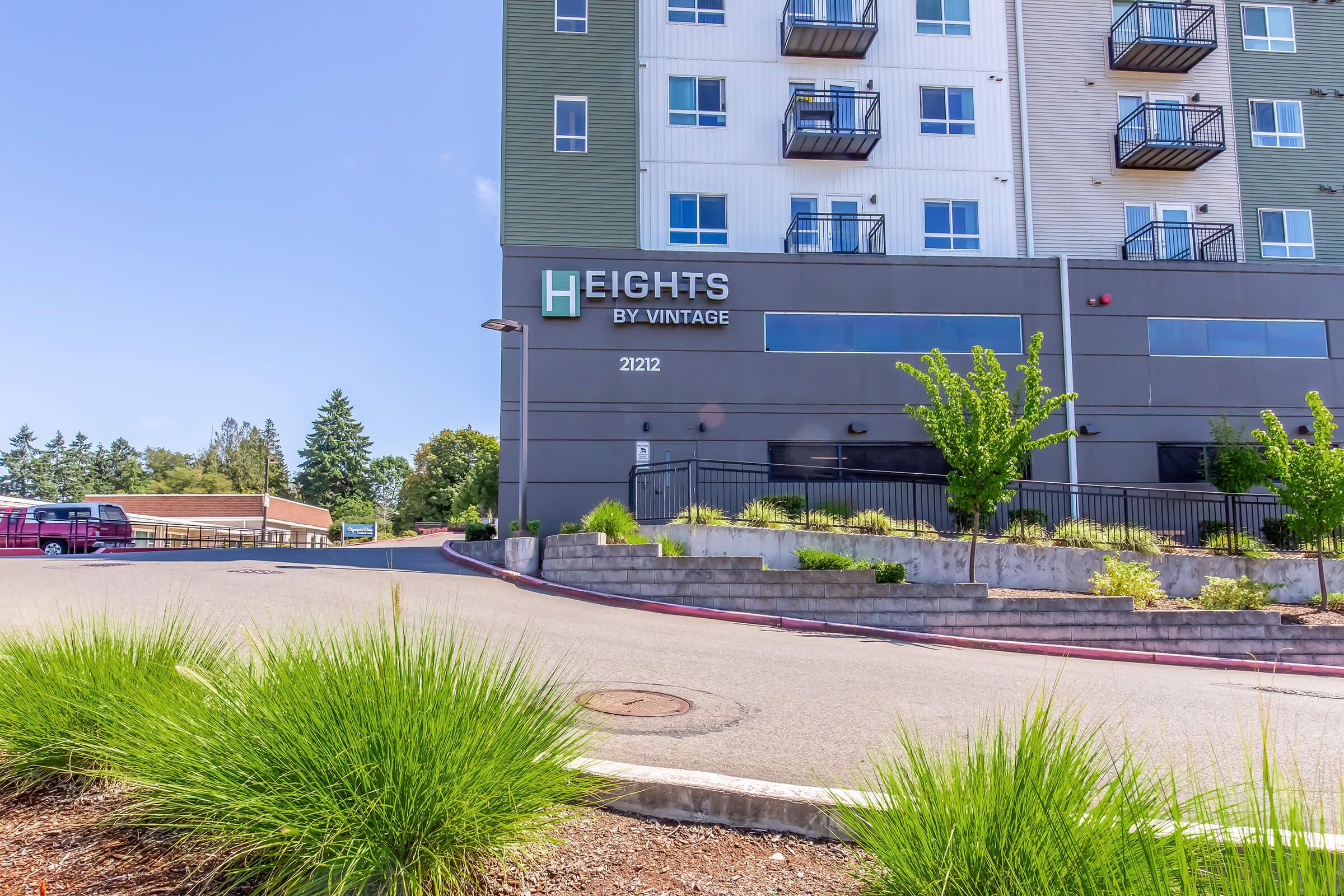 Modern apartment building with the sign "Heights by Vintage" visible. The building features multiple floors, balconies, and green landscaping in the foreground. A paved driveway leads up to the entrance, with trees and shrubs surrounding the area. Clear blue sky in the background.