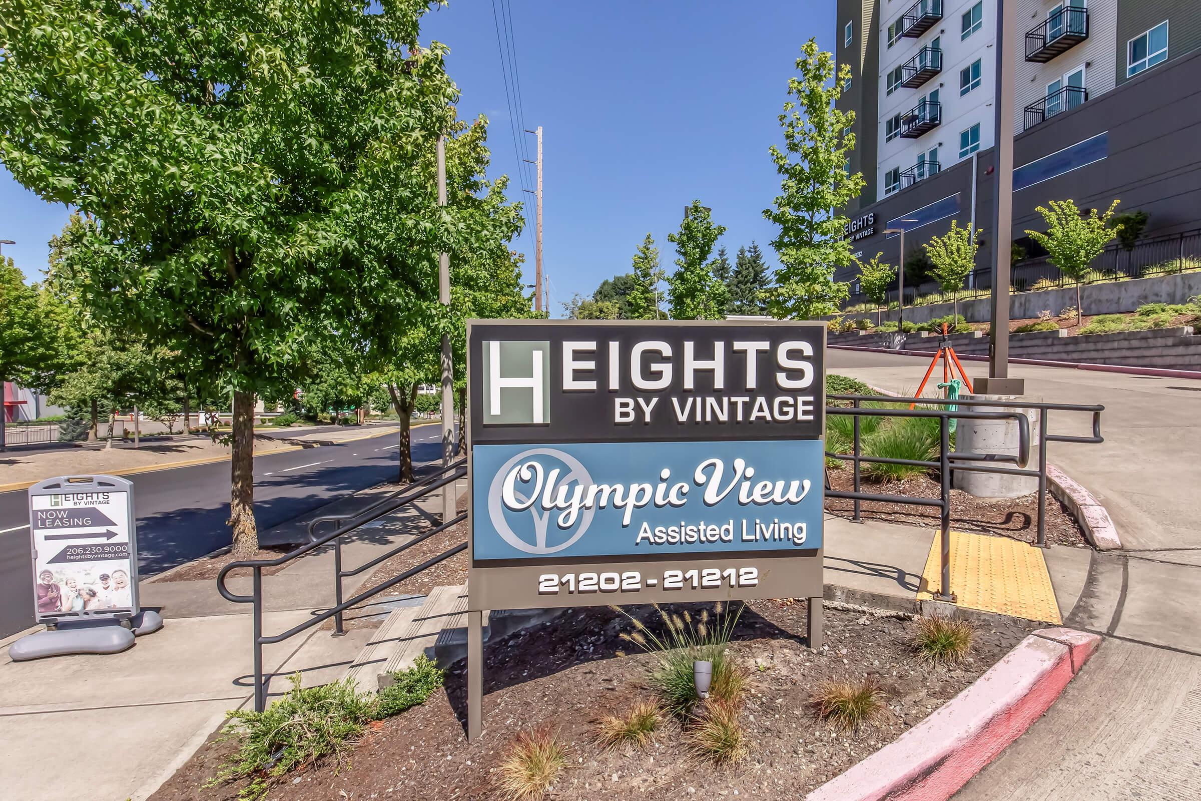 Sign for "Heights by Vintage" with "Olympic View Assisted Living" underneath. The image shows a clear, bright entrance with landscaped trees and a well-maintained pathway. The location is modern and inviting, suggesting a welcoming atmosphere for residents and visitors.