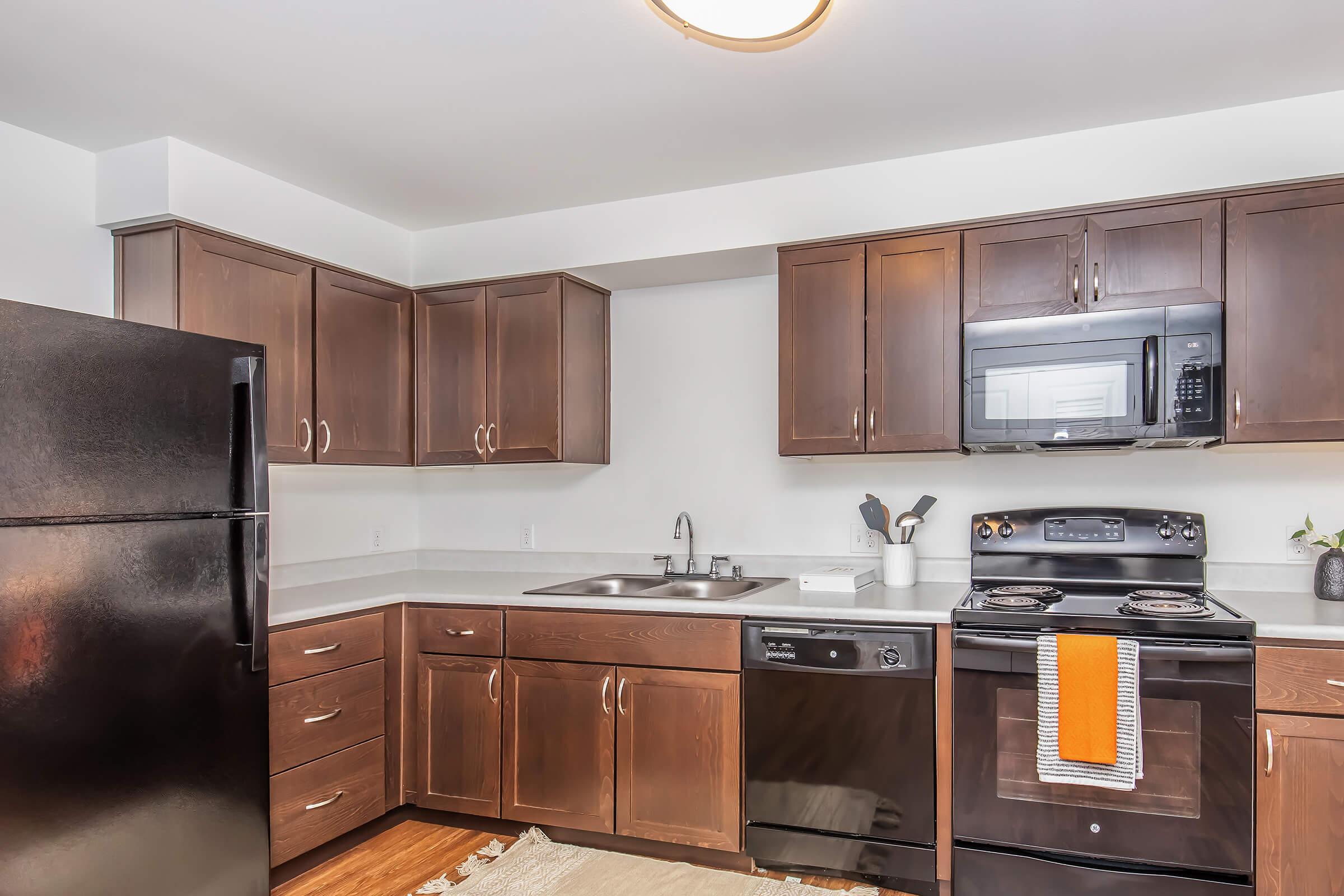 Modern kitchen featuring dark wooden cabinets, a stainless steel sink, black refrigerator, microwave, and oven. The countertop is light-colored, and there is a dishwasher under the counter. A kitchen utensil holder and an orange dish towel add a pop of color. The flooring is a warm wood tone.