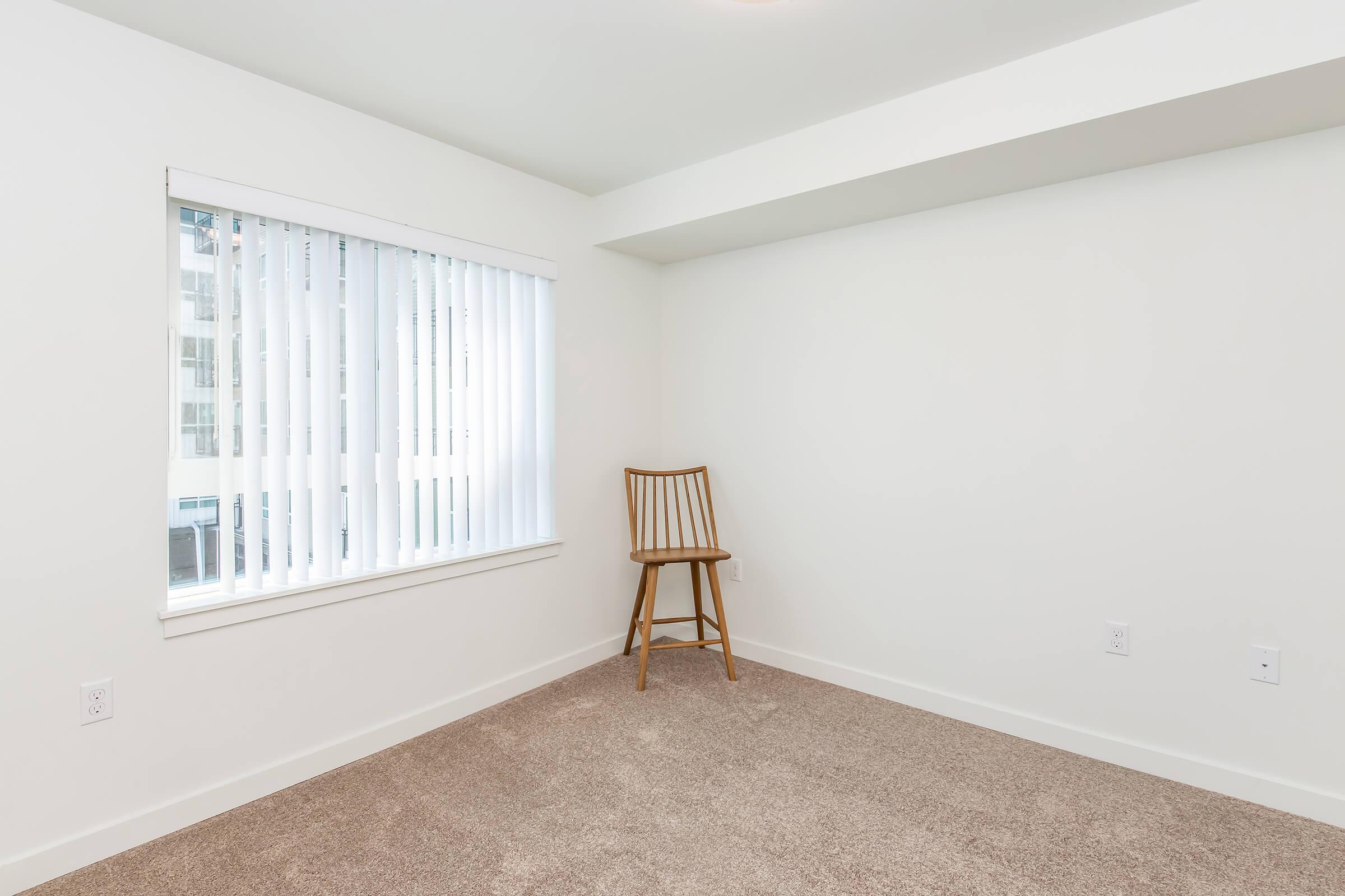 A simple, empty room with light beige carpet and white walls. A single wooden chair is positioned in the corner near a window with vertical blinds. The window allows natural light to enter, creating a bright and open feel in the otherwise minimalist space.