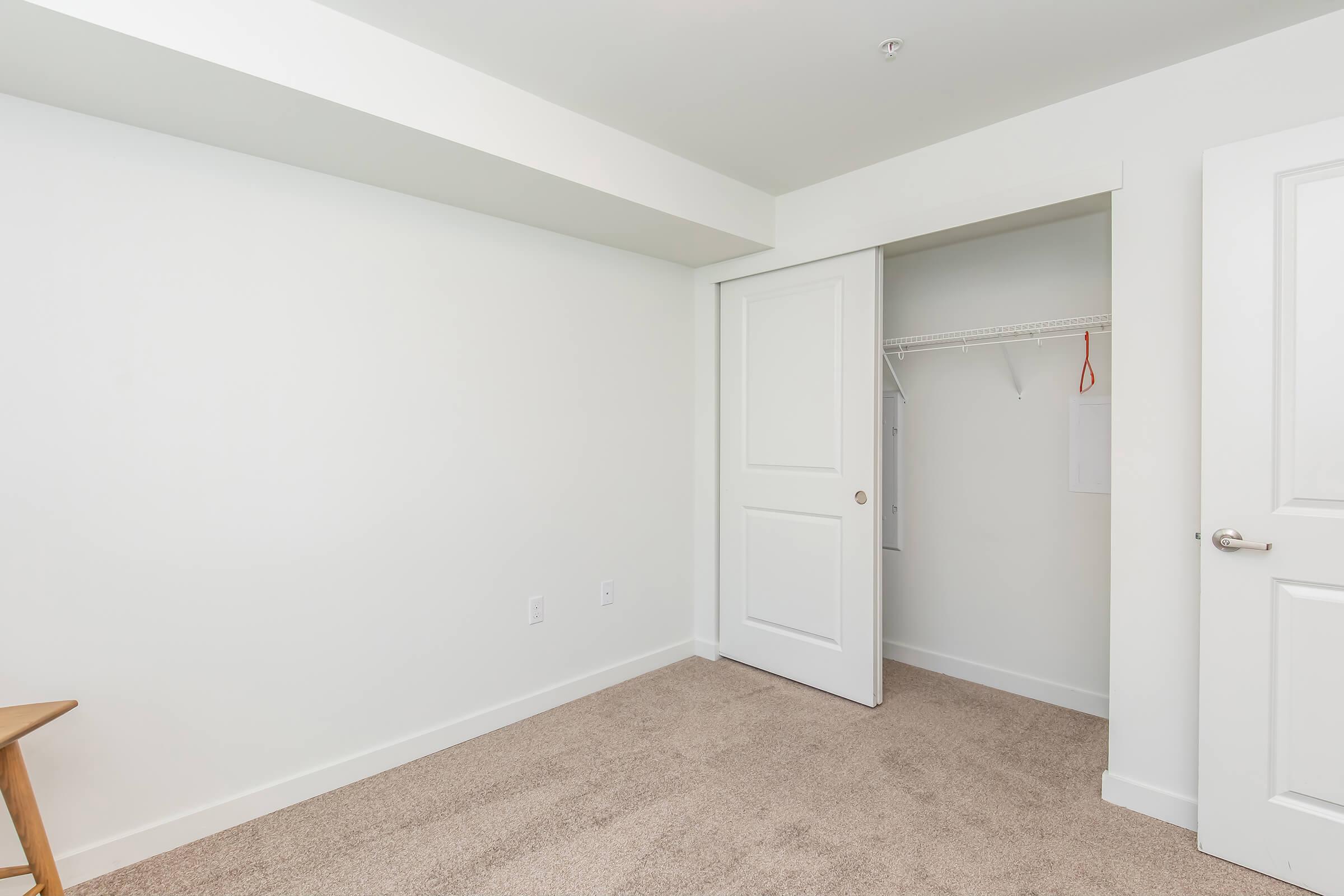 A vacant room with light-colored walls and carpet, featuring a closed white door leading to a closet with hanging space. There is a wooden chair in one corner, and the overall atmosphere is bright and minimalist, suggesting it could be a bedroom or office space.