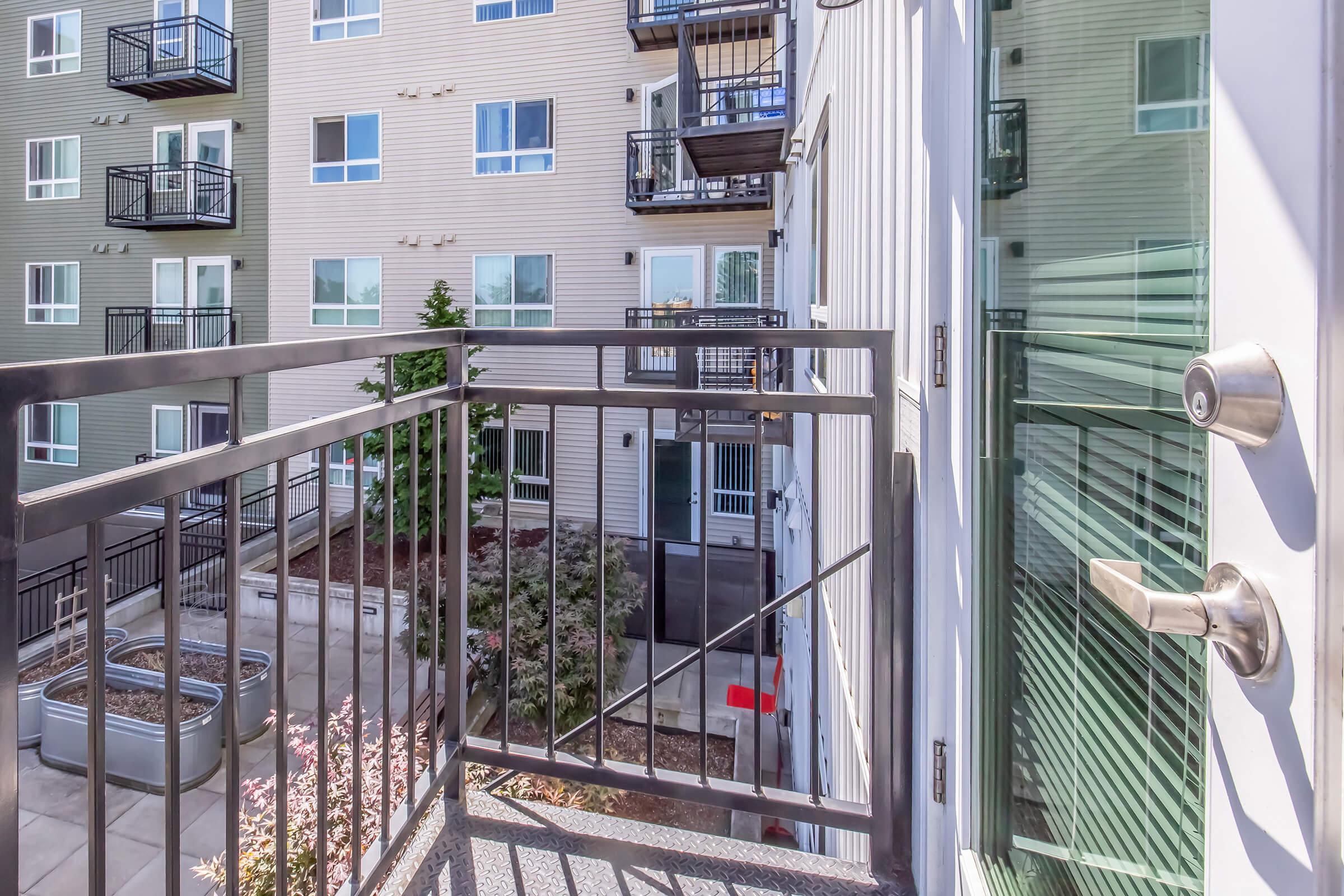 A small balcony with a metal railing overlooks a landscaped area and neighboring building. The balcony has a door leading into the apartment, and large windows reflect the nearby greenery and architecture. The scene emphasizes a modern urban living space.