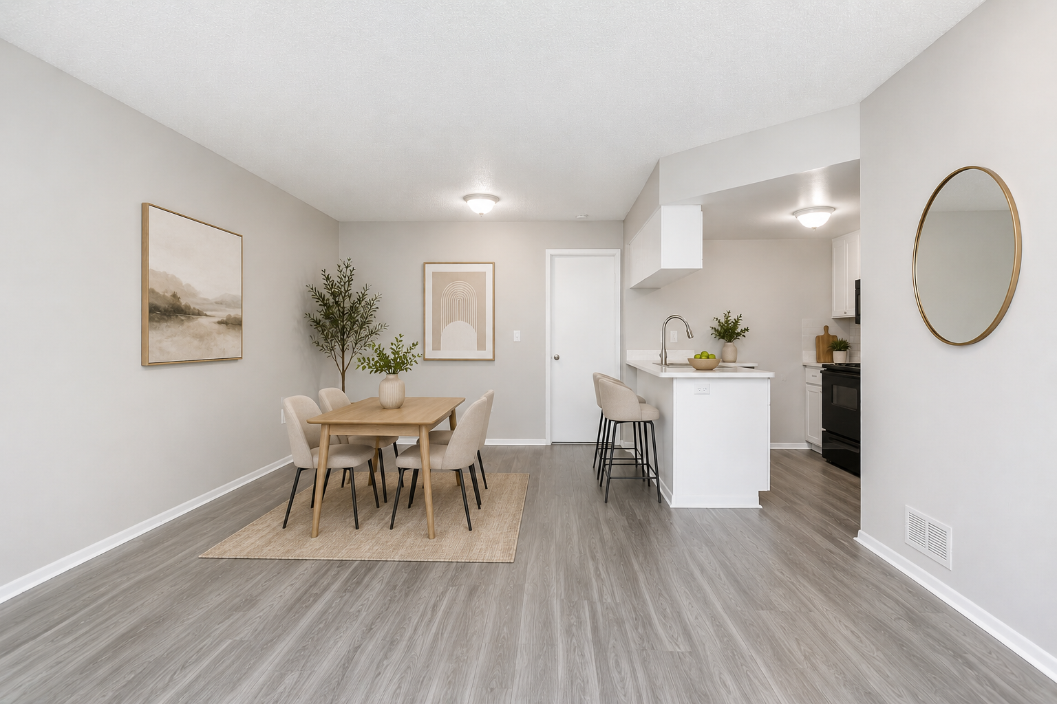 A modern, light-filled living space featuring a dining area with a round table and four chairs, a small kitchen island, and a minimalist design. The walls are painted in soft neutral tones, complemented by a piece of artwork and decorative plants. Natural light illuminates the area, enhancing the inviting atmosphere.
