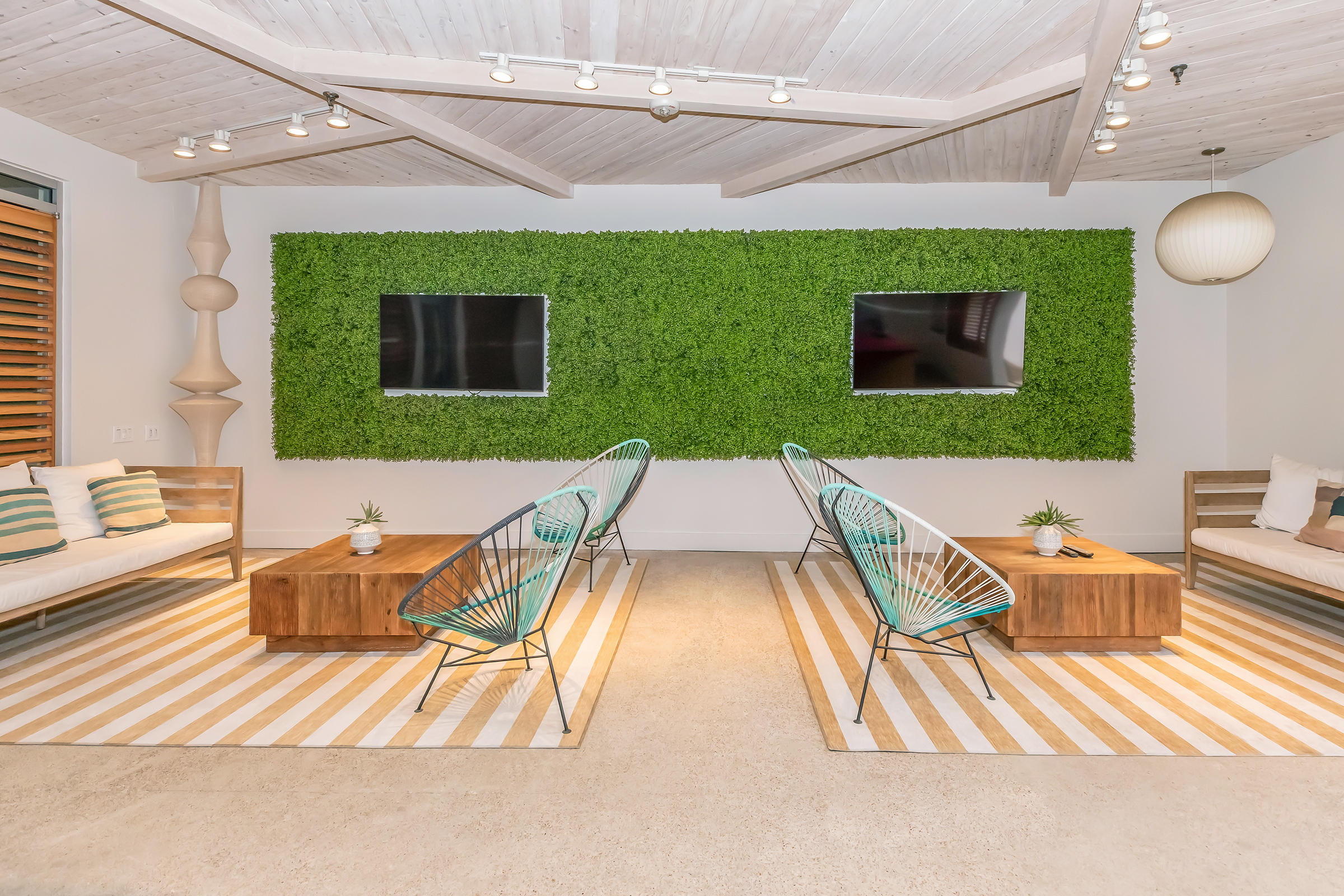 A modern lounge area featuring two wall-mounted televisions above a vibrant green moss wall. There are two wooden coffee tables and two contemporary chairs, with beige sofas on either side. The room has a light-colored concrete floor and is decorated with striped rugs, creating a stylish and inviting atmosphere.