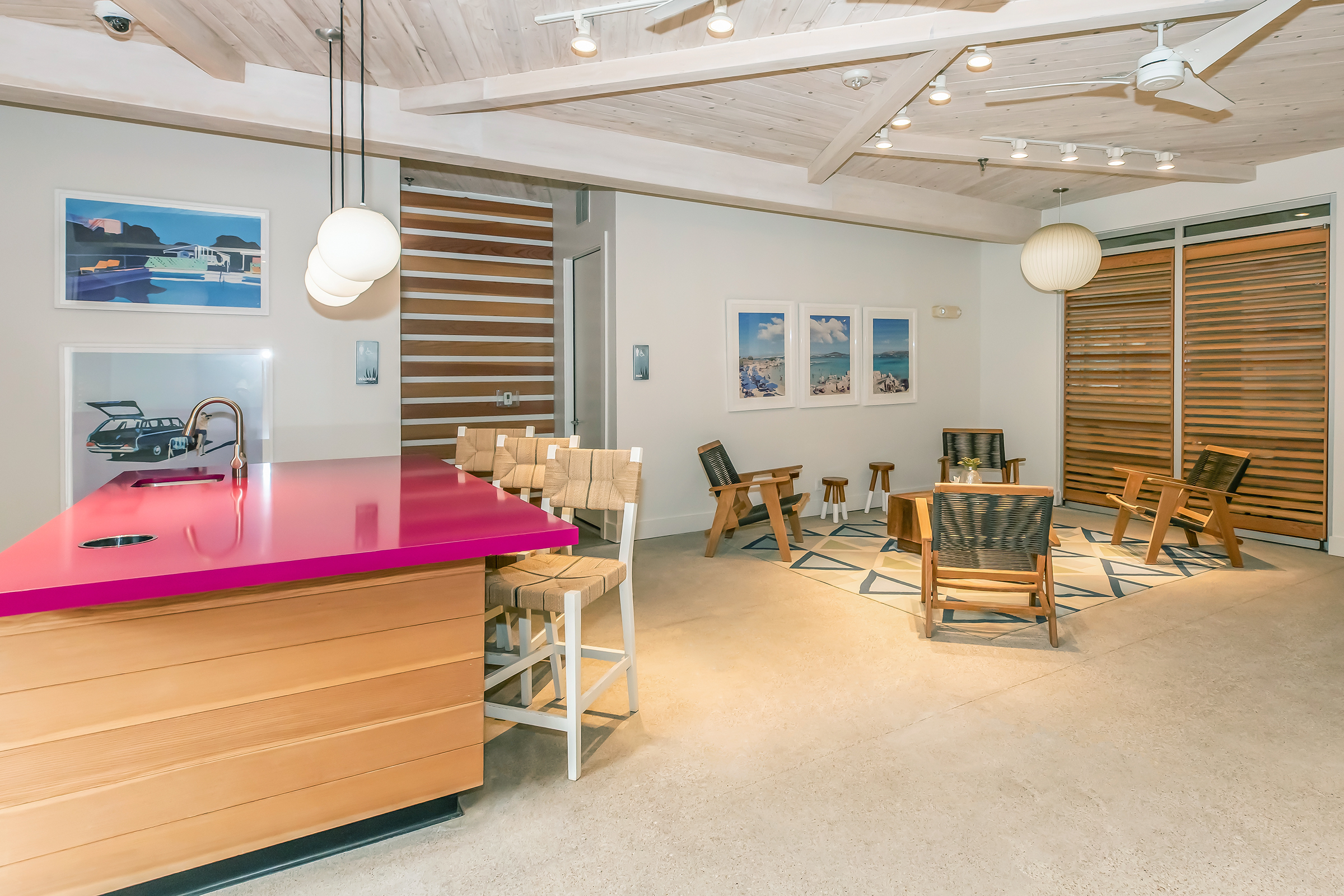 Brightly lit modern lounge area featuring a pink countertop bar with stools, wooden chairs arranged in a casual seating area, and a stylish wood-paneled wall. The space has artwork on the walls and unique light fixtures, creating a welcoming and contemporary atmosphere.
