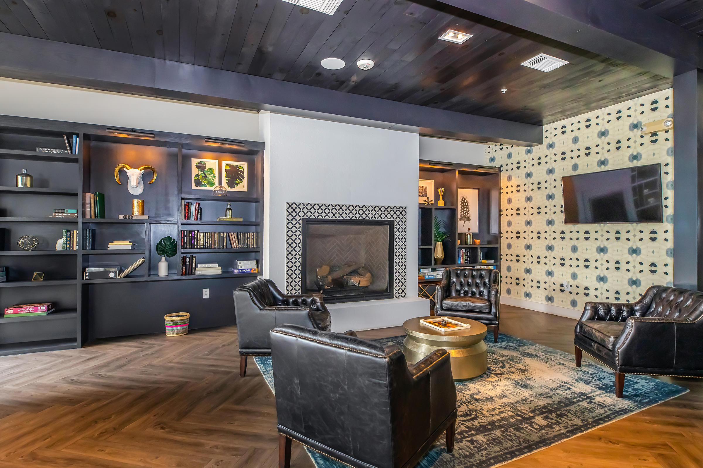 A stylish living room featuring a modern fireplace, two black leather chairs, and a gold coffee table. The walls are decorated with a patterned wallpaper, and there are shelves filled with books and decorative items. A television is mounted on one wall, creating a cozy and inviting atmosphere.