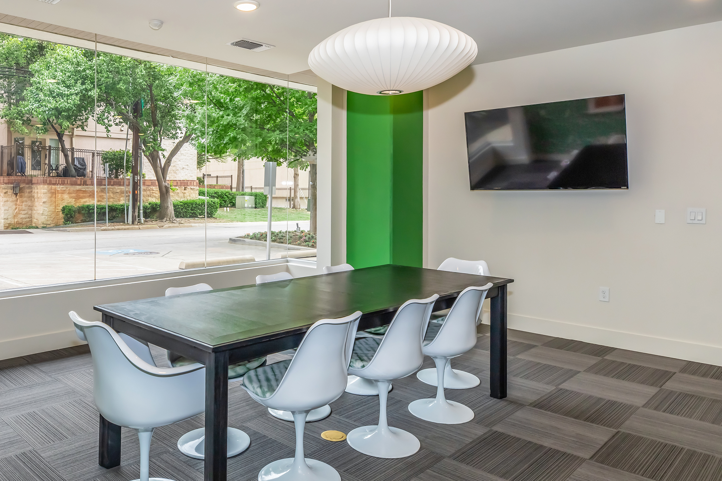 A modern meeting room featuring a long black table surrounded by white swivel chairs. In the background, large windows provide natural light and view of greenery outside. A flat-screen TV is mounted on the wall, and there is a unique pendant lamp hanging from the ceiling, creating a bright and inviting atmosphere.