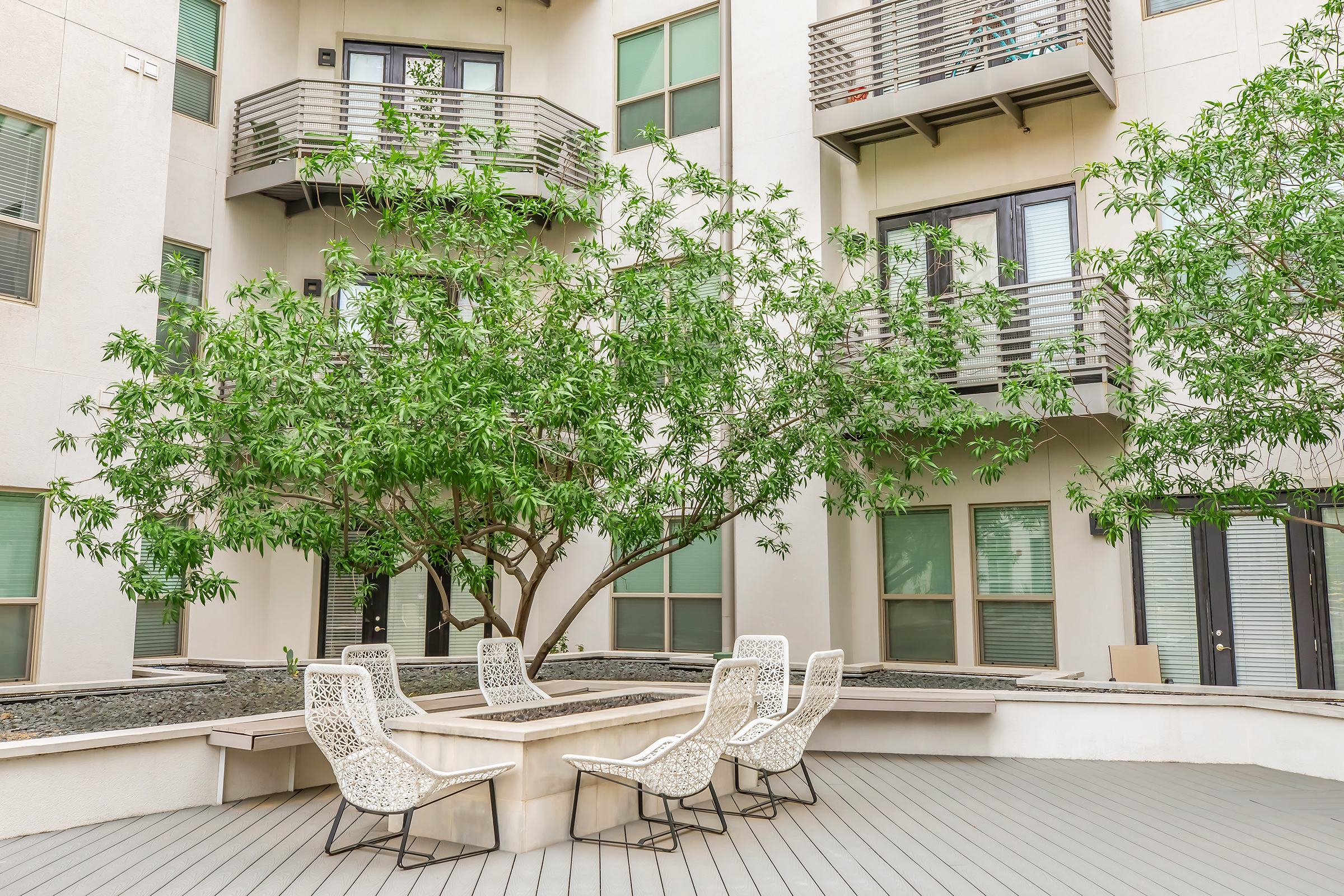 A modern outdoor patio area featuring several white chairs arranged around a small table. Lush greenery from nearby trees and plants enhances the space, while a view of apartment windows and balconies in the background adds to the urban ambiance. The wooden deck provides a warm, inviting setting.
