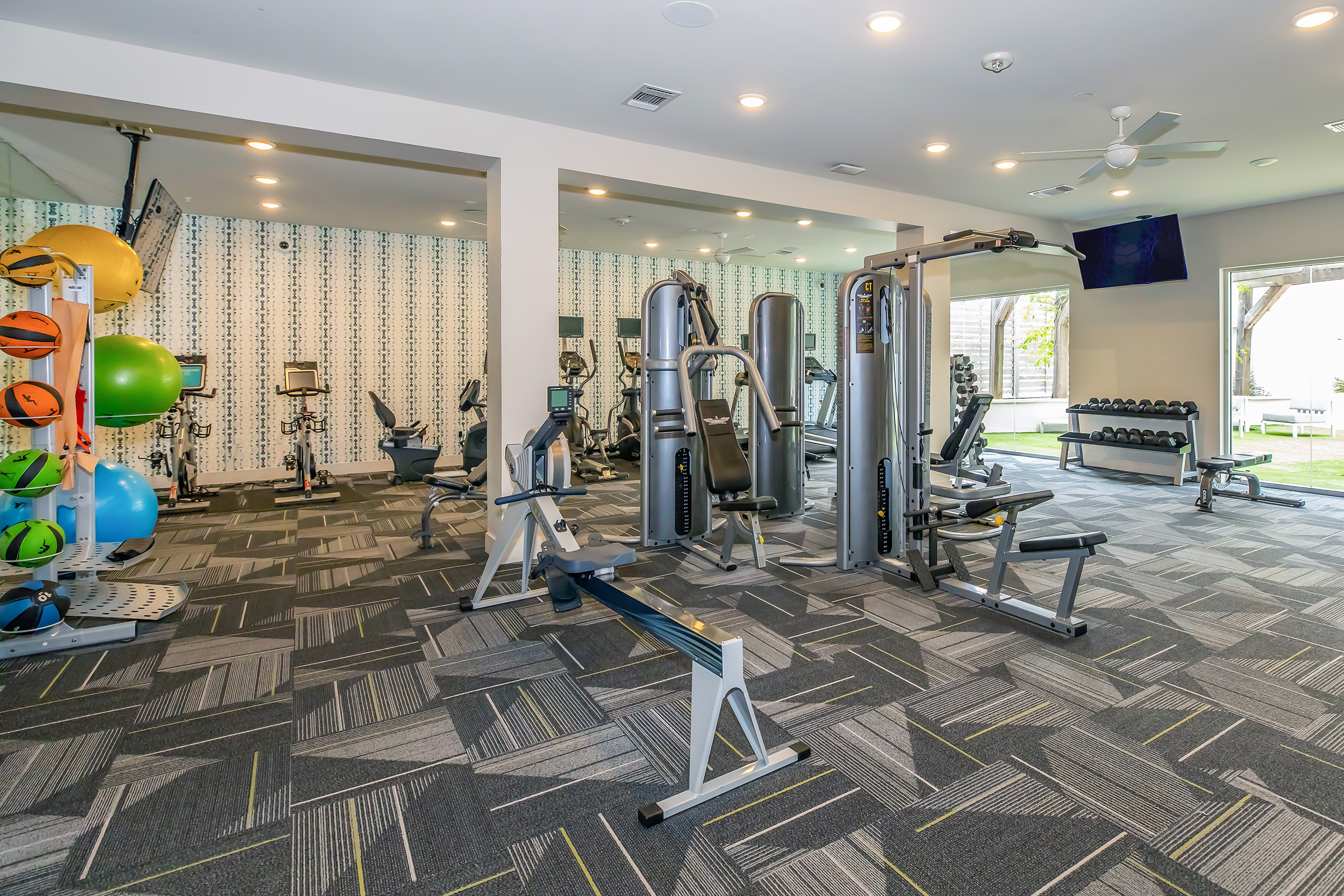 A modern gym interior featuring various exercise equipment, including weight machines, free weights, and cardio machines. The space is well-lit and has a stylish design with patterned walls and colorful fitness balls. Large windows allow natural light, enhancing the inviting atmosphere for workouts.