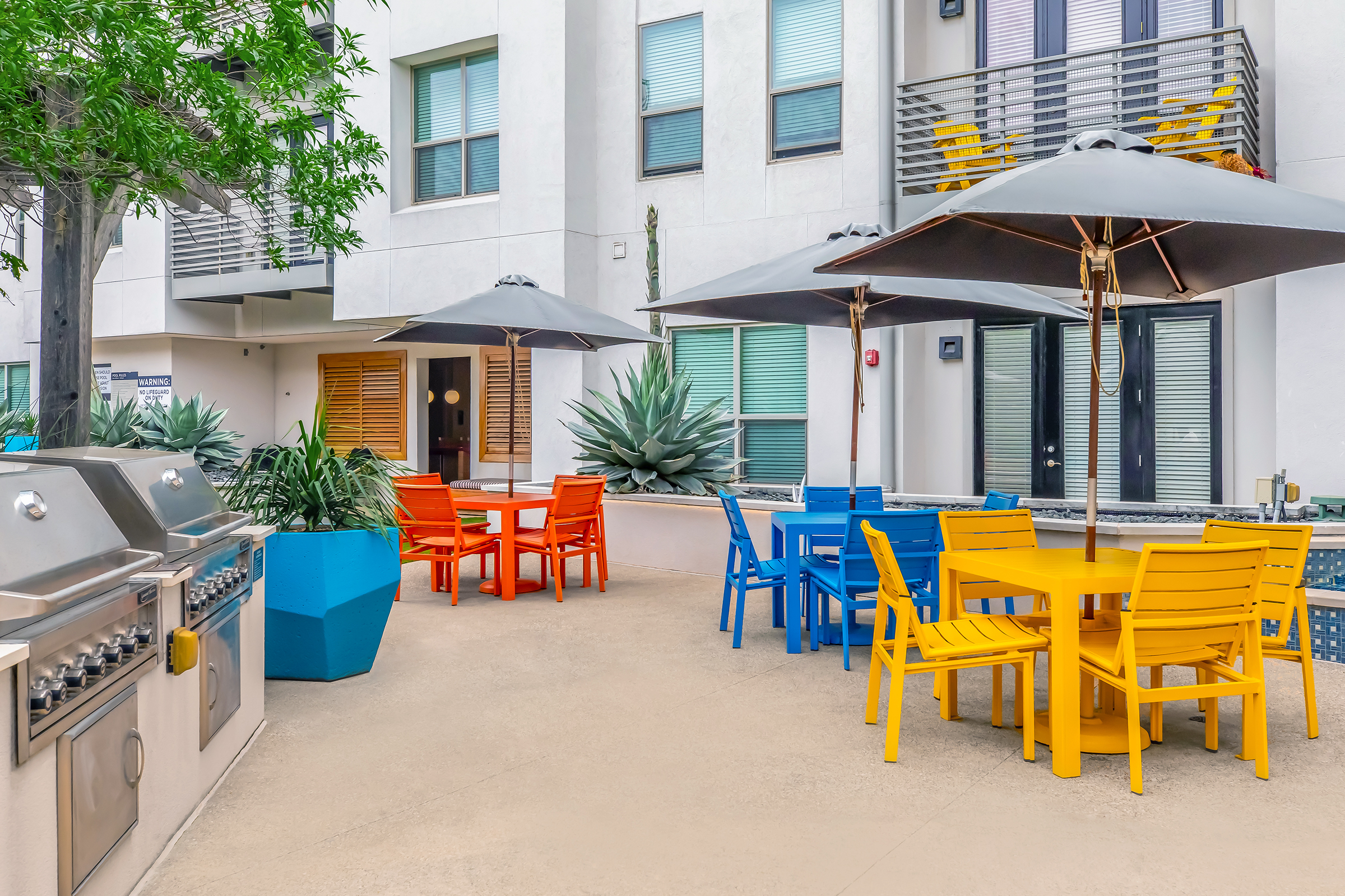 Outdoor patio area featuring colorful furniture with yellow and blue tables and chairs, large umbrellas providing shade, and a grill station. Lush plants and a decorative agave plant add greenery, against a backdrop of modern, white buildings with large windows.
