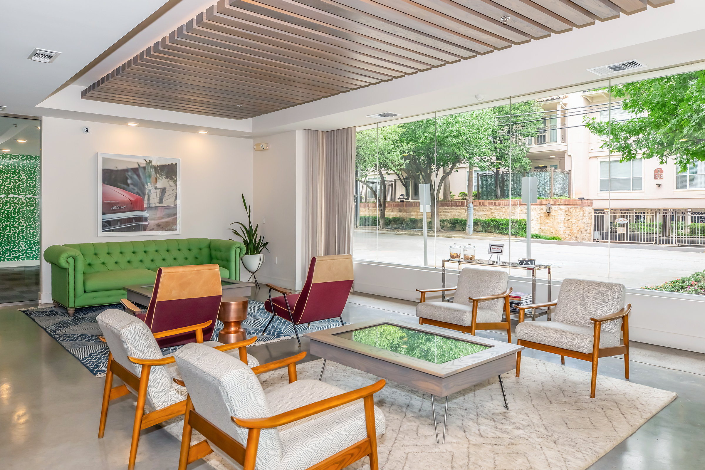 Modern lounge area featuring a green sofa, two red armchairs, and light-colored wooden chairs. A coffee table sits on a textured rug, and a large window provides a view of trees and an outdoor space. The room has a contemporary design with wooden ceiling panels and minimalist decor.