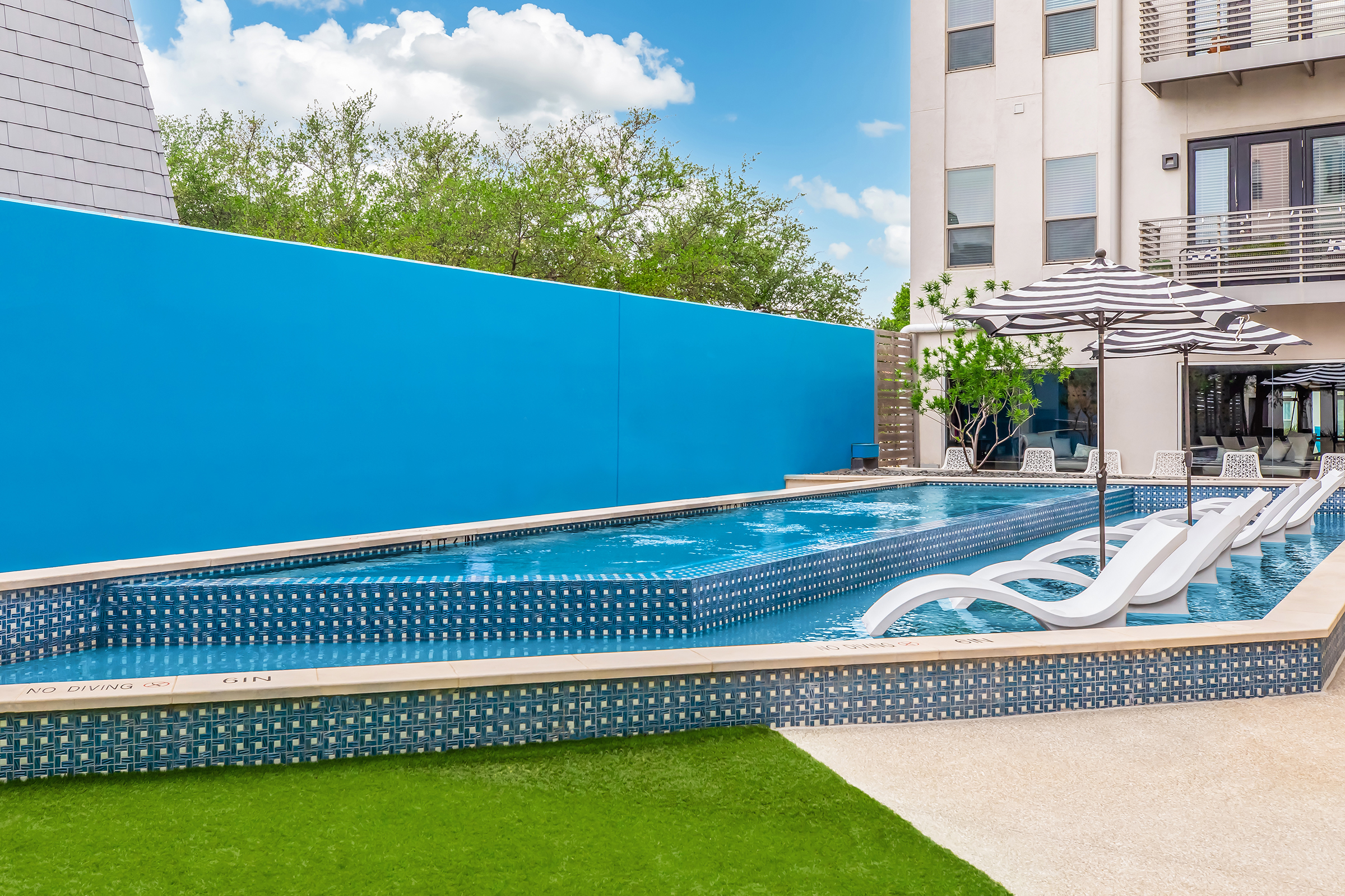 A modern outdoor swimming pool surrounded by a vibrant blue wall. Lounge chairs with white frames are positioned alongside the pool, which features a striking tile design. Lush greenery and a clear blue sky complete the relaxing atmosphere, ideal for leisure and outdoor enjoyment.