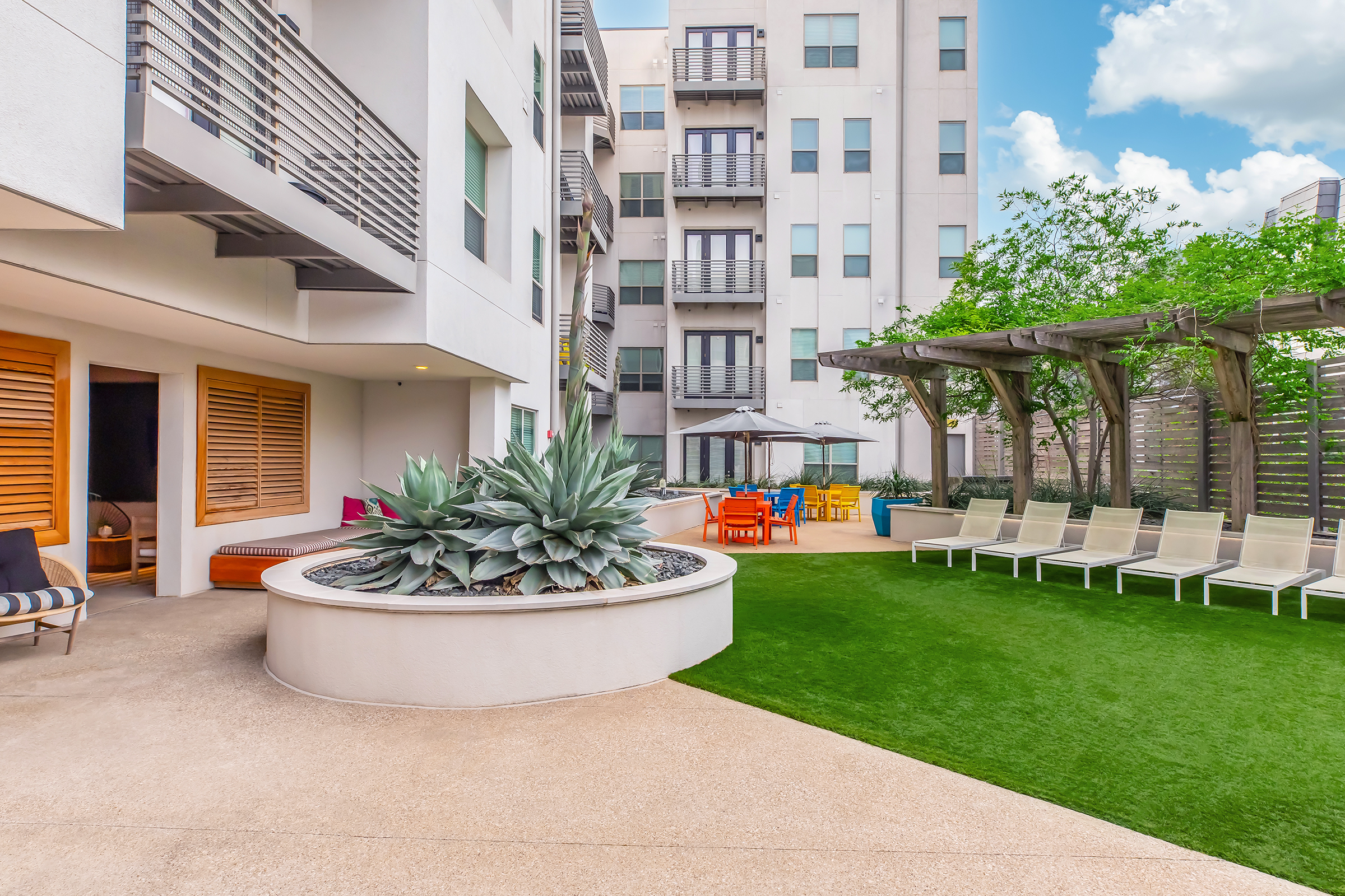 A modern outdoor space featuring a landscaped garden with neatly trimmed grass, seating areas, lounge chairs, and a pergola. The scene includes apartment buildings in the background, along with vibrant patio furniture, creating a welcoming and relaxing atmosphere. Clouds are visible in a bright blue sky.