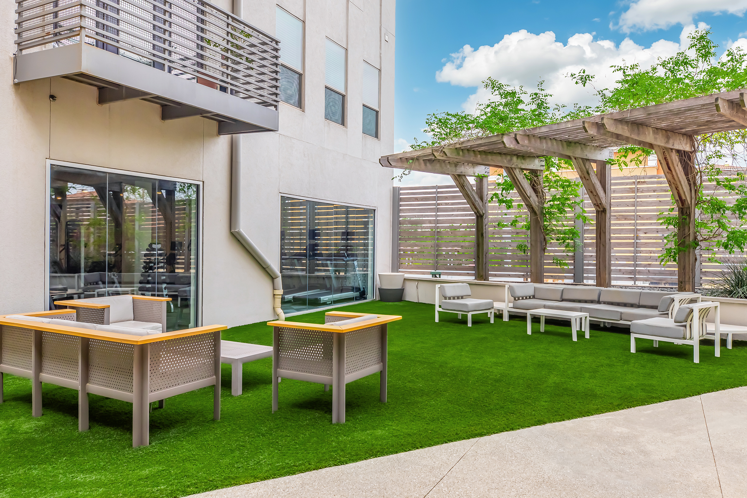 A modern outdoor space featuring artificial grass, comfortable seating areas with gray and white furniture, a wooden pergola, and a glass wall. The setting is bright and inviting, with natural light filtering through nearby greenery and a clear blue sky above.