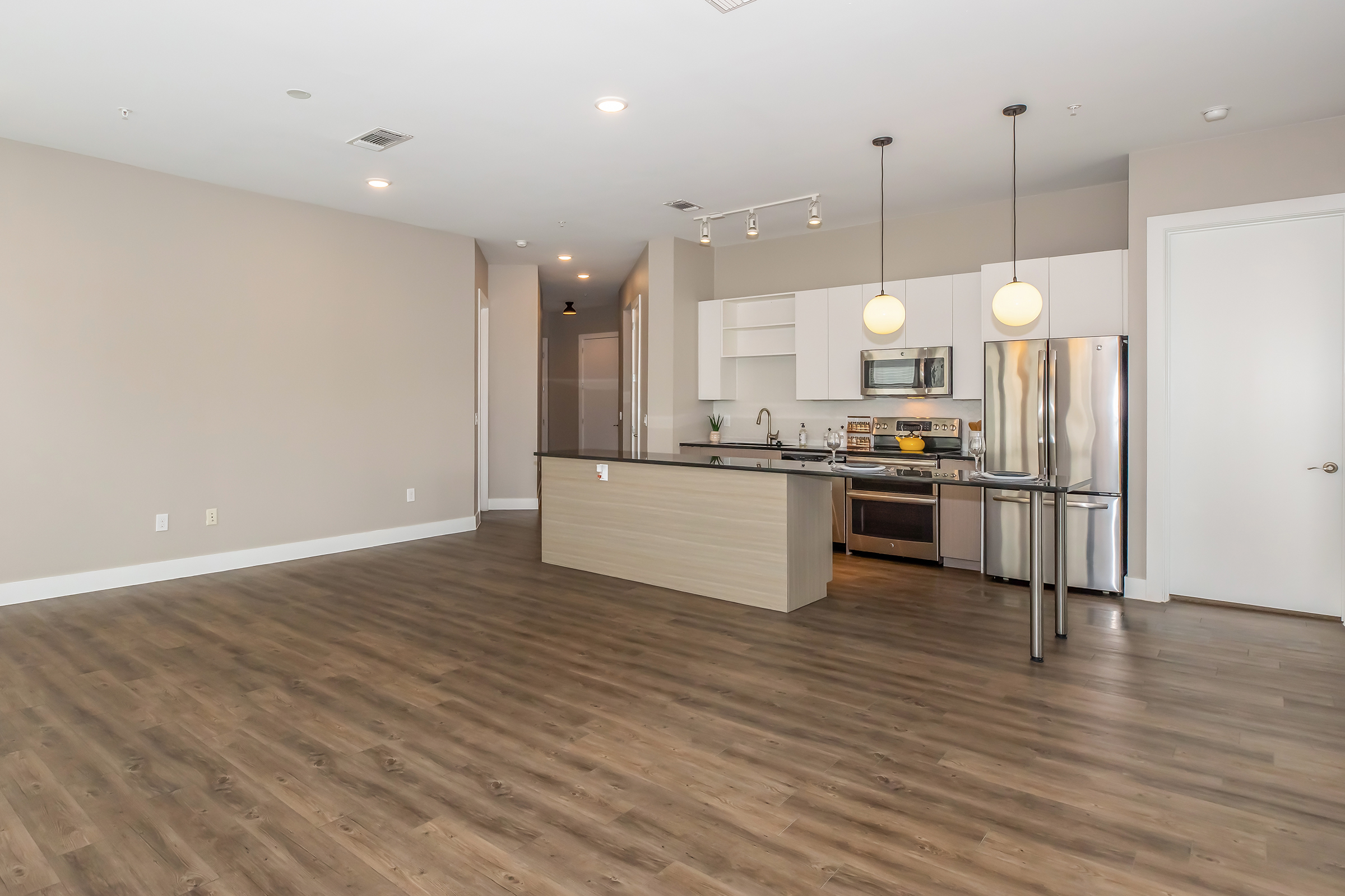 A modern, open-concept kitchen and living area featuring light brown wood flooring, white walls, and minimalistic decor. The kitchen includes stainless steel appliances, an island with seating, and pendant lighting. Natural light fills the space, creating a bright and inviting atmosphere.