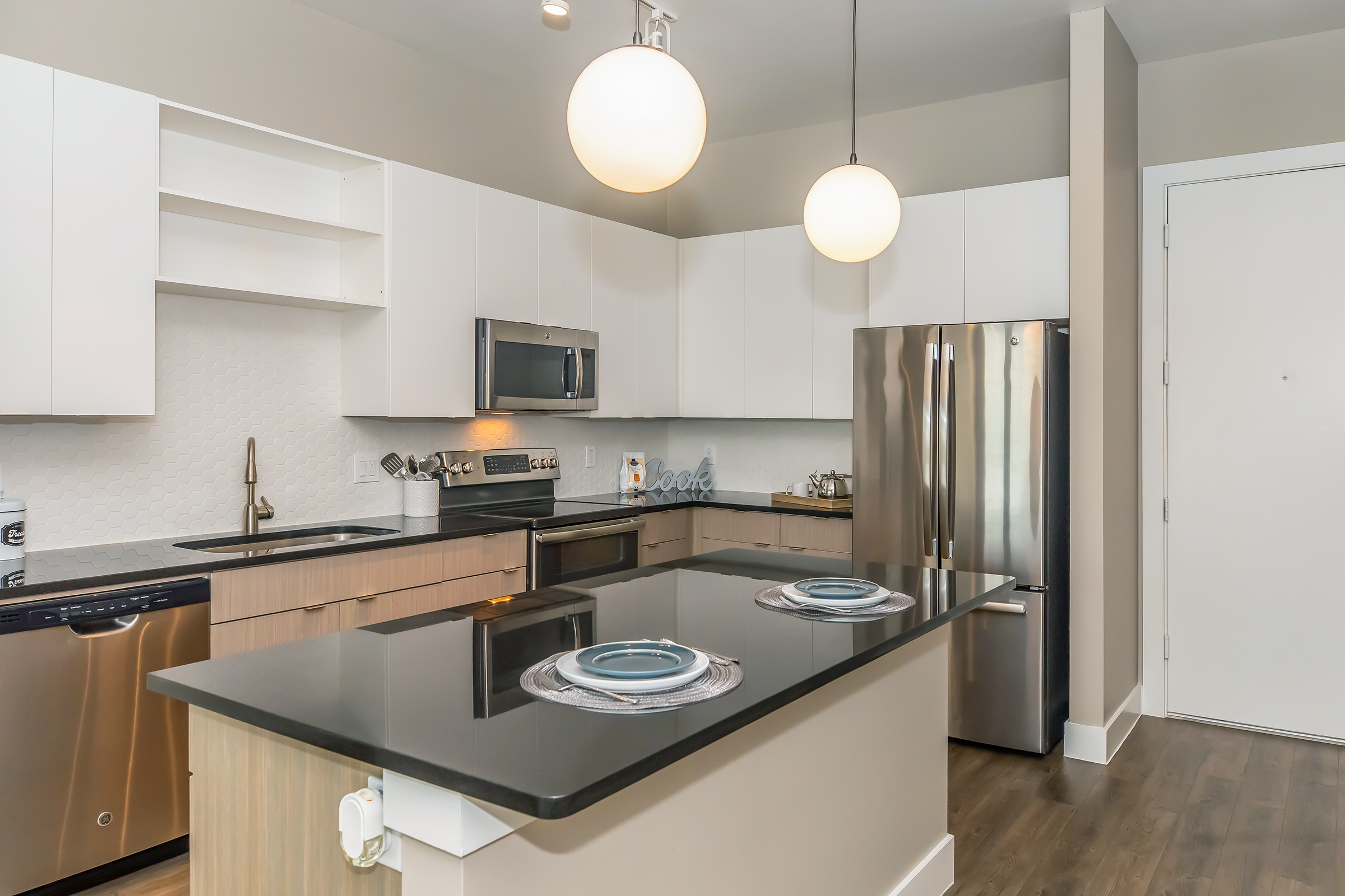 Modern kitchen featuring a central island with a dark granite countertop, two pendant lights overhead, white cabinetry, and stainless steel appliances including a fridge and oven. The kitchen has a clean, minimalist design with wooden flooring and a contemporary aesthetic.