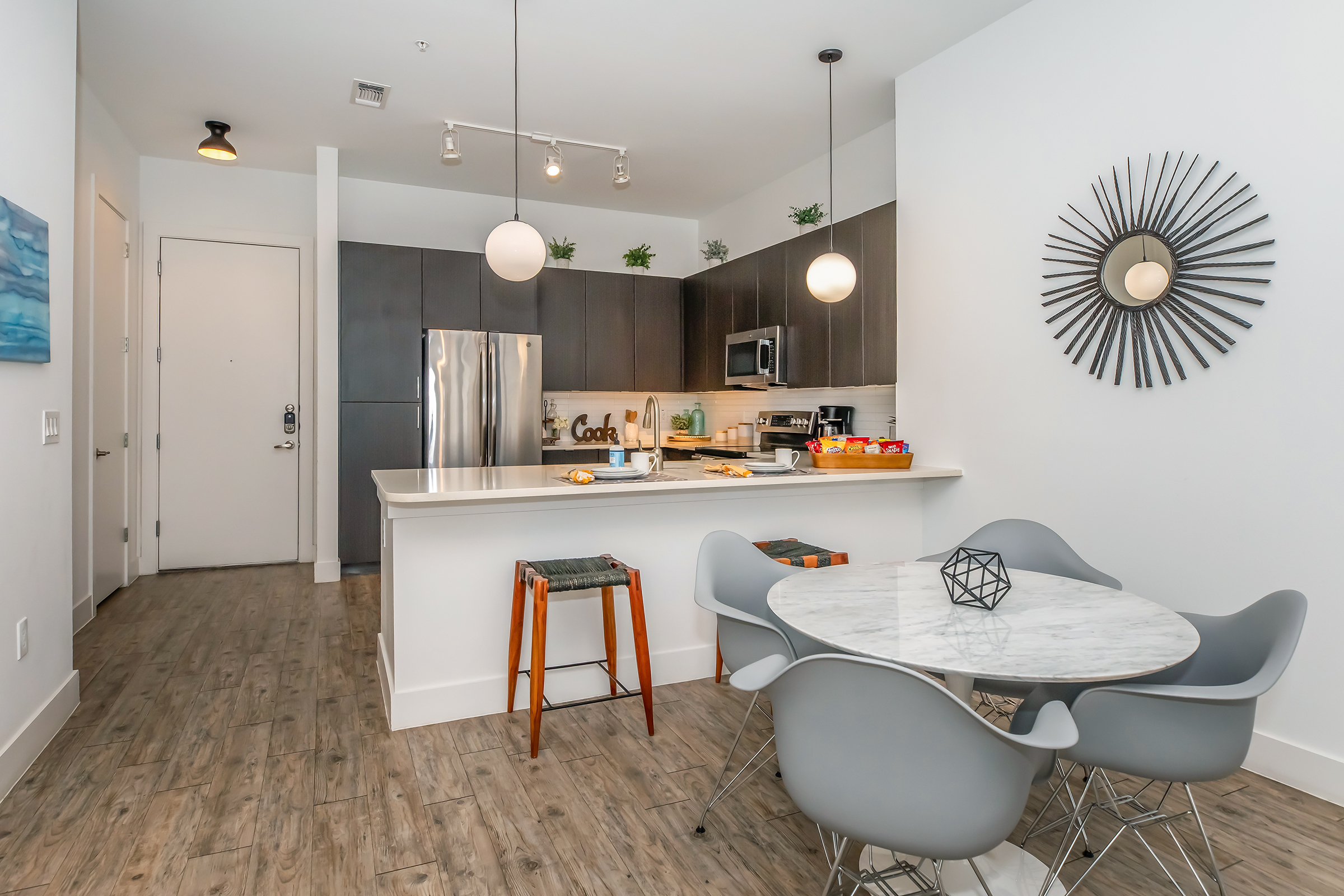 Modern kitchen and dining area featuring dark cabinets, stainless steel appliances, and a white countertop. A round marble dining table with gray chairs is set in the foreground. The space includes decorative elements like a wall mirror and plants, along with a door leading outside. Bright, contemporary design with wooden flooring.
