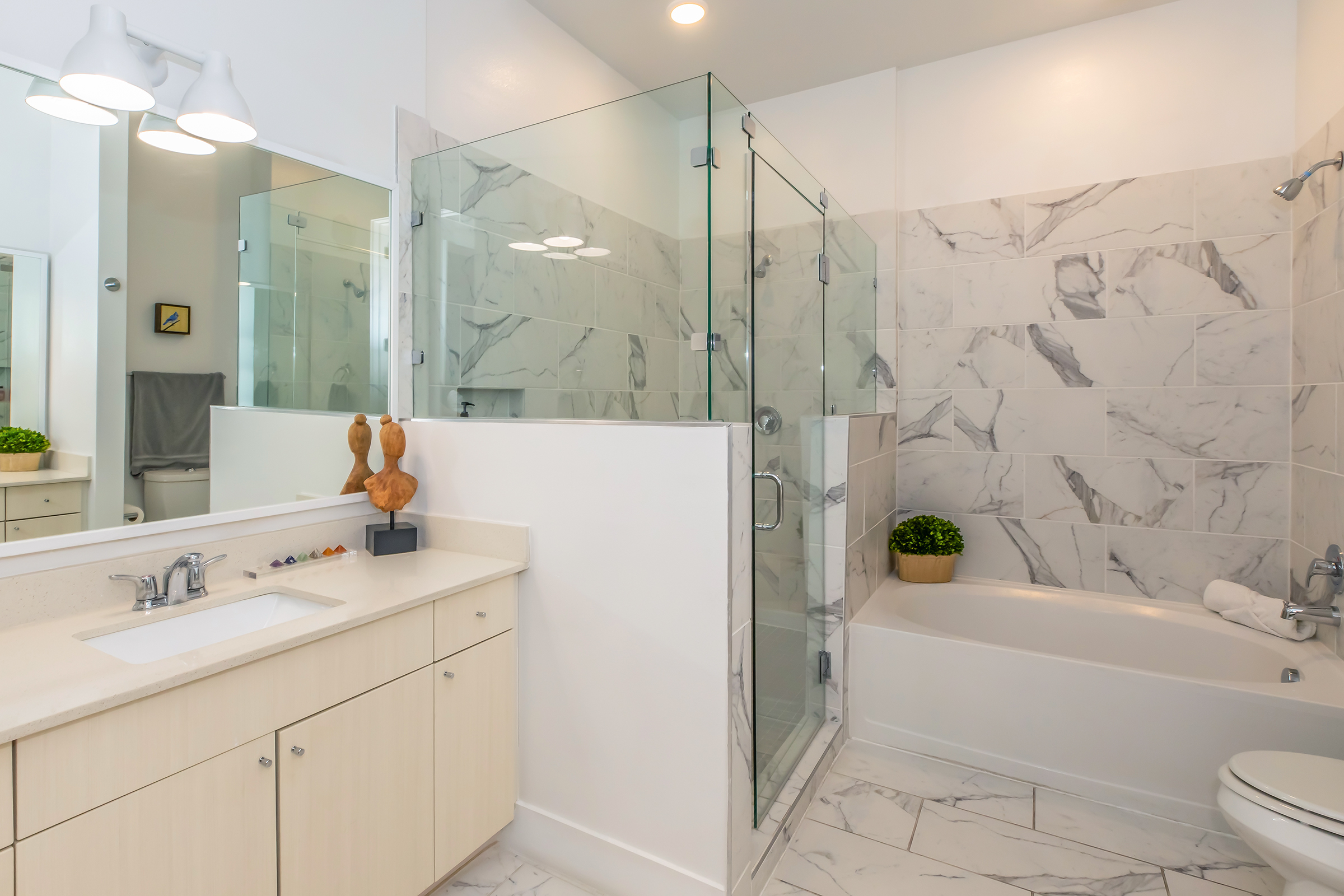 Modern bathroom featuring a glass shower enclosure, a bathtub, and a minimalist vanity with wooden accents. Light marble tiles adorn the walls, complemented by sleek fixtures and bright lighting. A small potted plant adds a touch of greenery, enhancing the contemporary aesthetic.