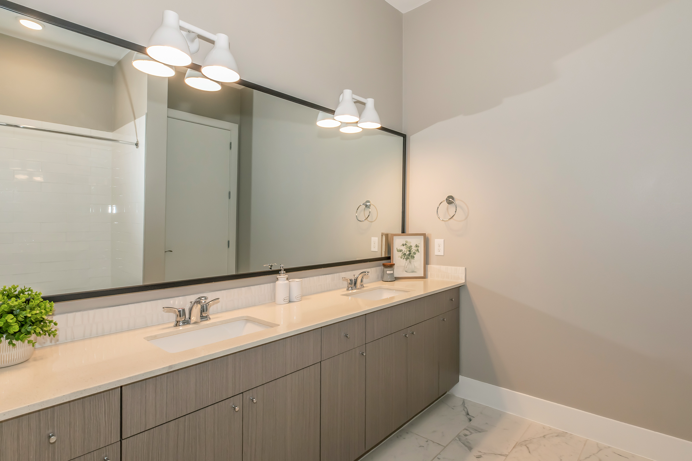 A modern bathroom featuring a double sink vanity with a light-colored countertop and gray cabinetry. Above the sinks, there are two large mirrors and stylish wall-mounted lamps. The walls are painted in a soft gray, and a small potted plant adds a touch of greenery. The floor is covered in white and gray marble tiles.
