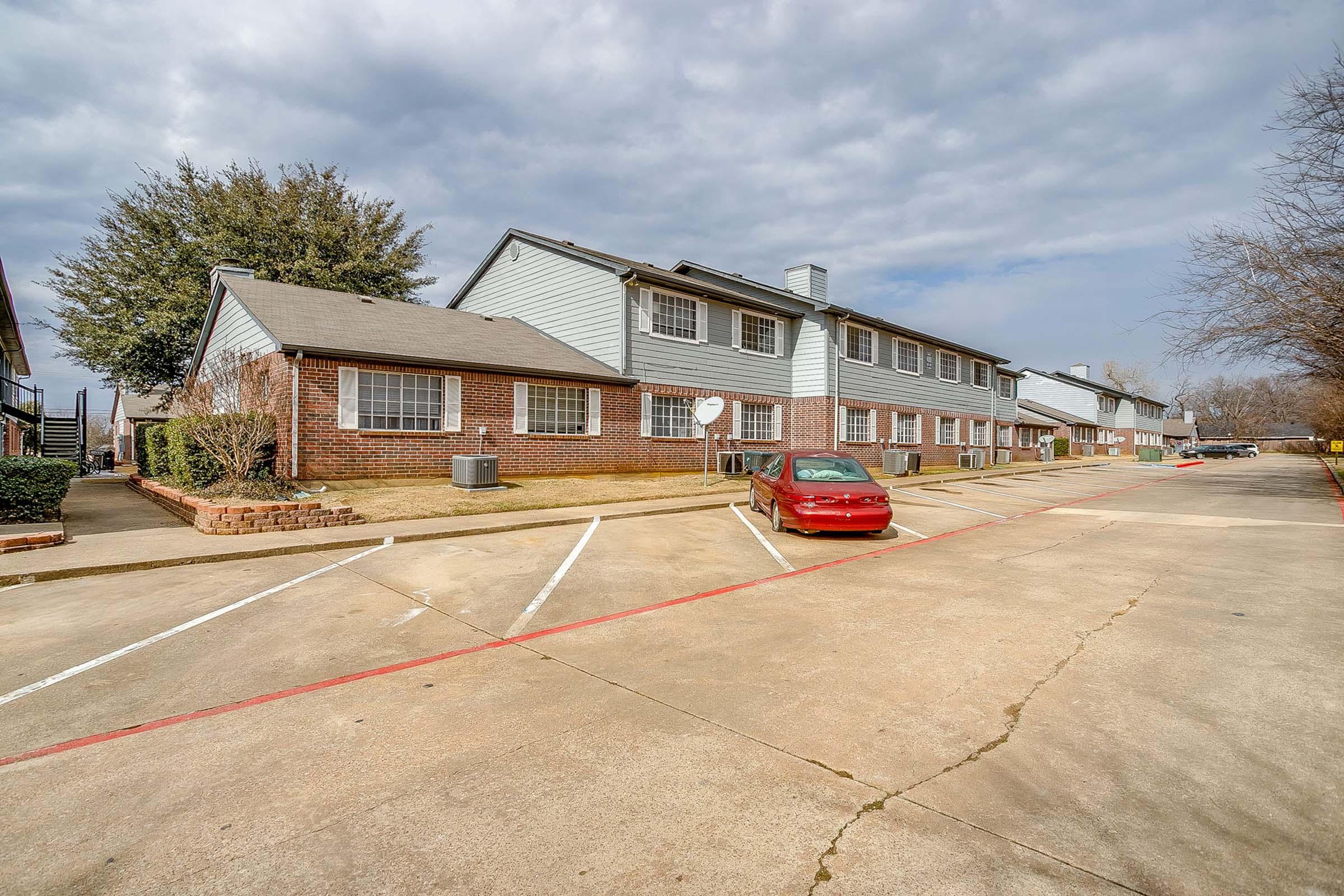 A wide view of a two-story apartment building with brick and siding exterior. The building is set in a parking lot with marked spaces, including a red car parked in front. The sky is cloudy, and there are trees visible near the building, adding to the residential atmosphere.