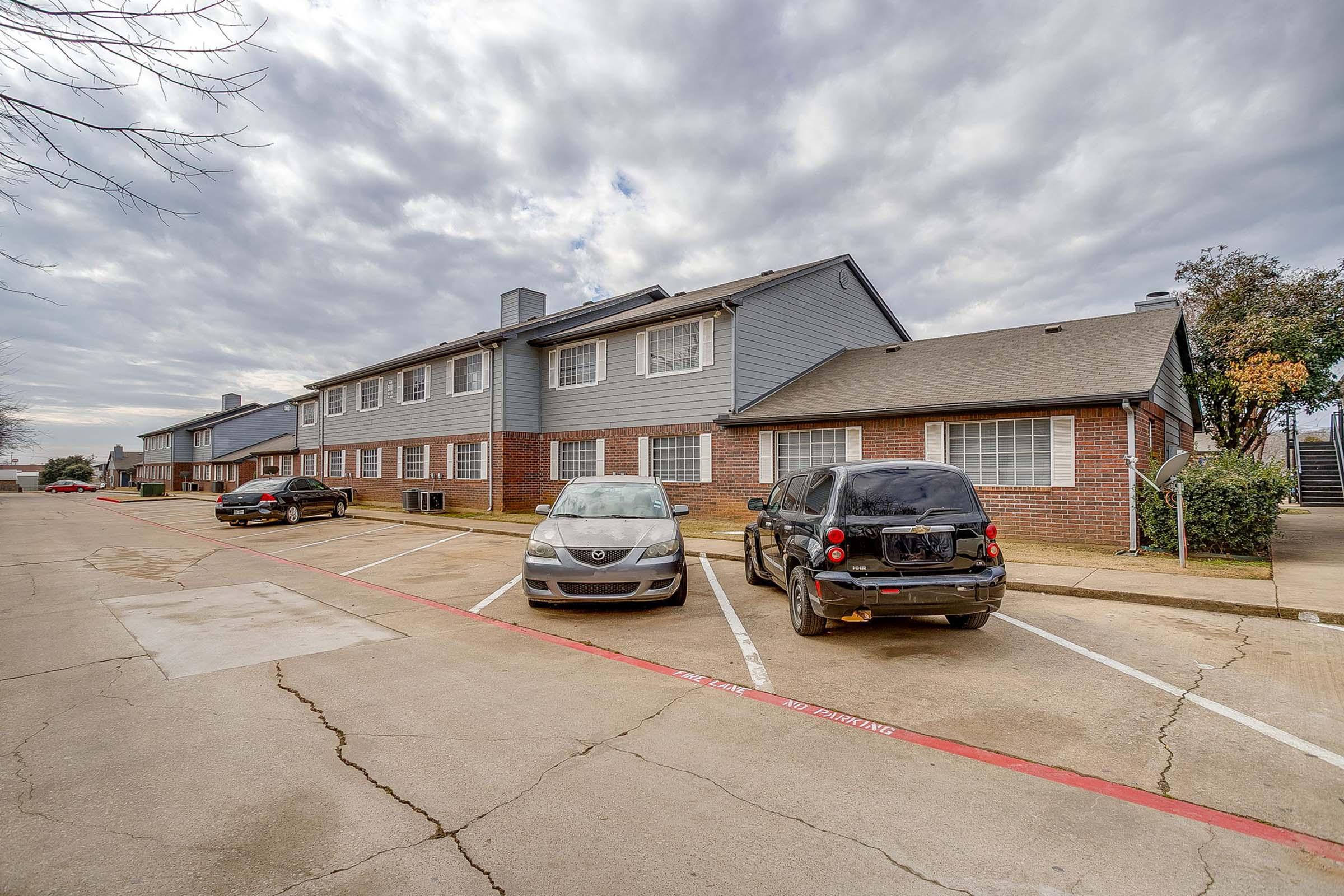 A residential apartment complex featuring two-story brick buildings under a cloudy sky. Two parked cars are visible in the foreground, with a red painted line marking parking spaces. The area appears well-maintained with green shrubs nearby.