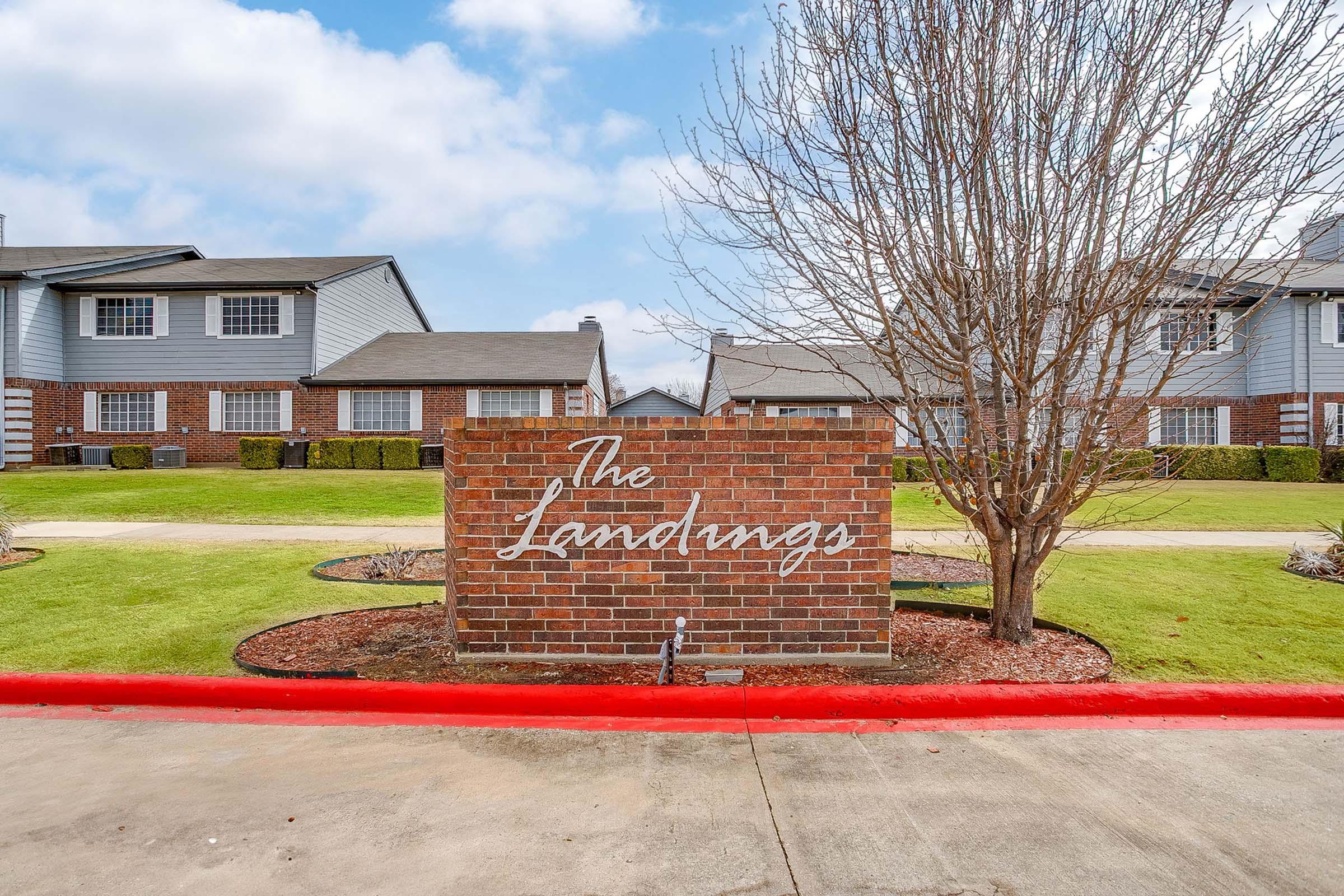 Sign for "The Landings" community, set against a well-maintained landscape featuring green grass and a backdrop of residential buildings. The sign is made of brick with white lettering, and there are bare trees nearby under a partly cloudy sky. A red curb runs along the front.