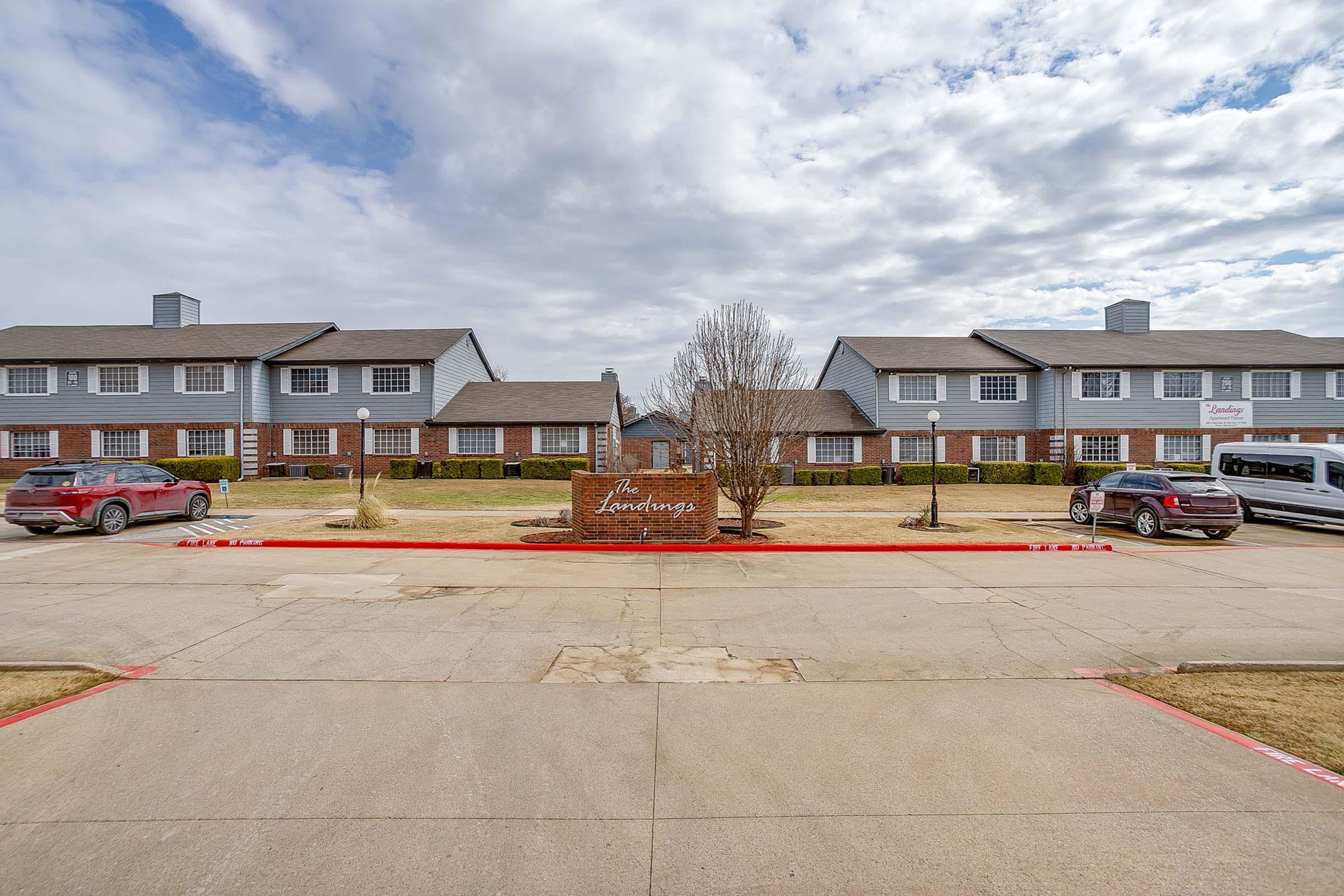 A view of a residential complex named "The Laramie," featuring two buildings with a landscaped area in front. The scene includes parking spaces with vehicles and a cloudy sky overhead. Decorative lighting fixtures are visible along the pathway.