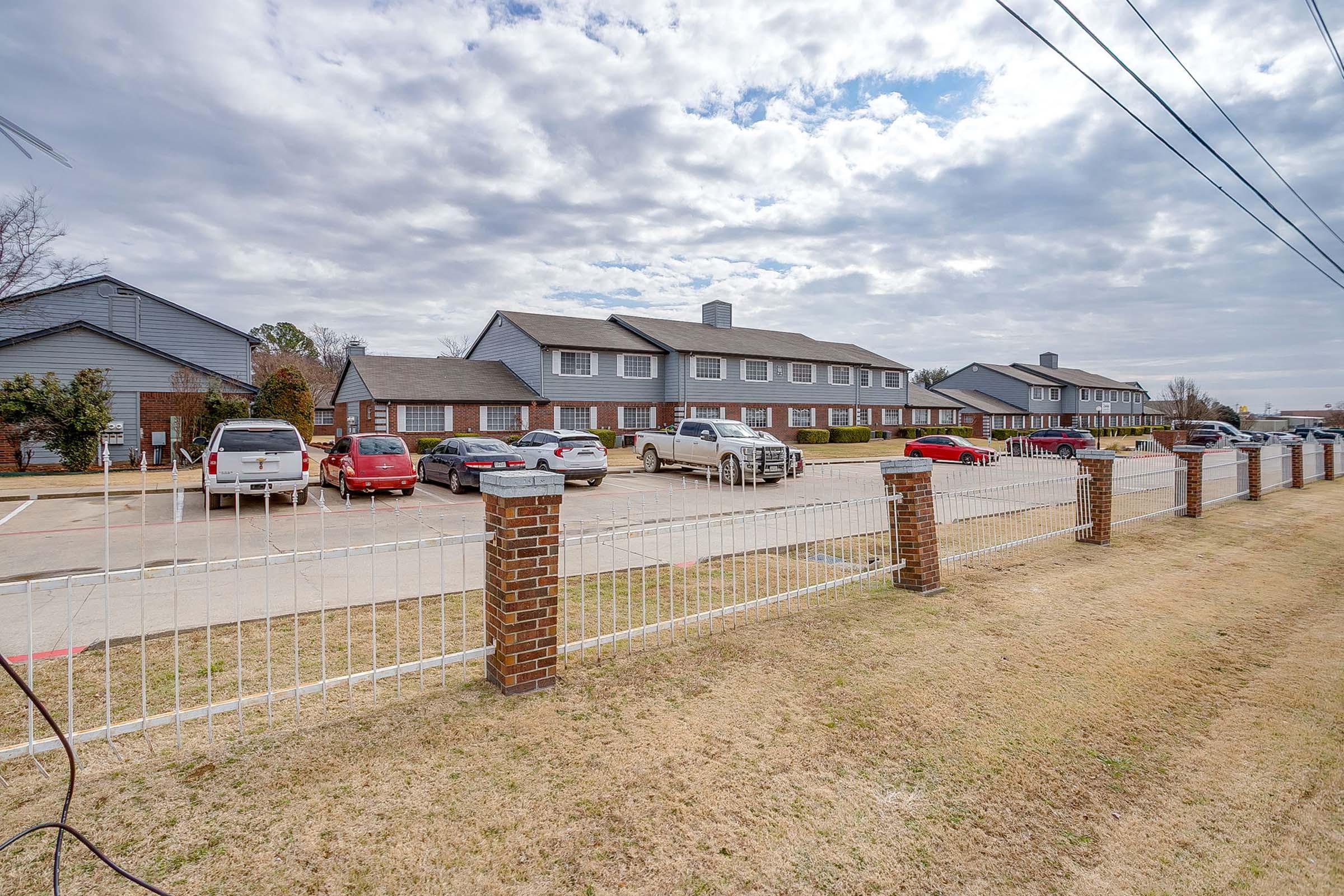 A row of multi-unit residential buildings with a parking area in front. Several cars are parked in the lot, and there's a grassy area with a low brick fence along the street. The sky is partly cloudy, indicating a mild day. Utility lines are visible in the foreground.