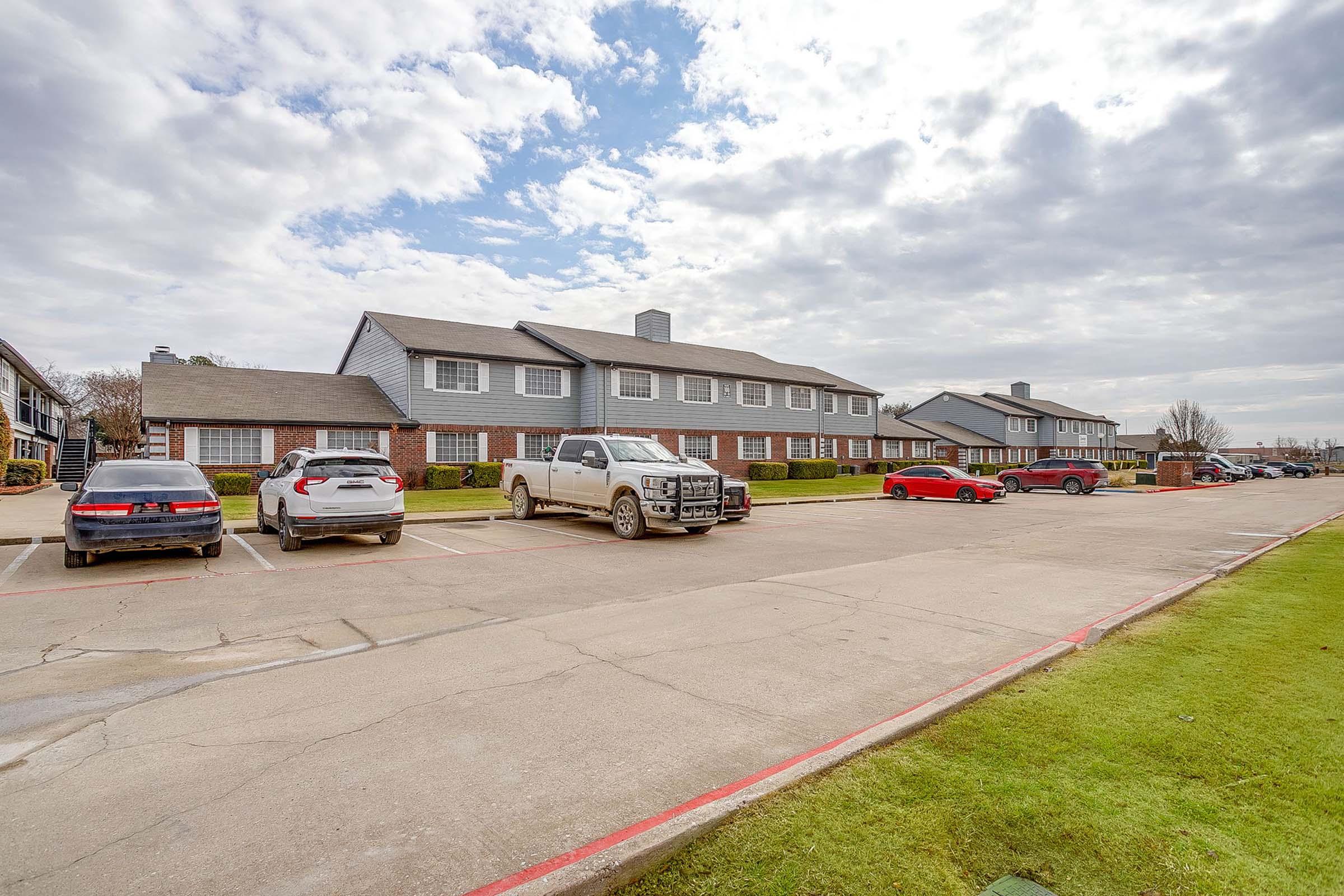 A parking lot with several cars, including a red sedan, a white SUV, a gray truck, and two additional sedans. In the background, there are two rows of two-story apartment buildings under a partly cloudy sky. Grass runs alongside the parking area, creating a green space.