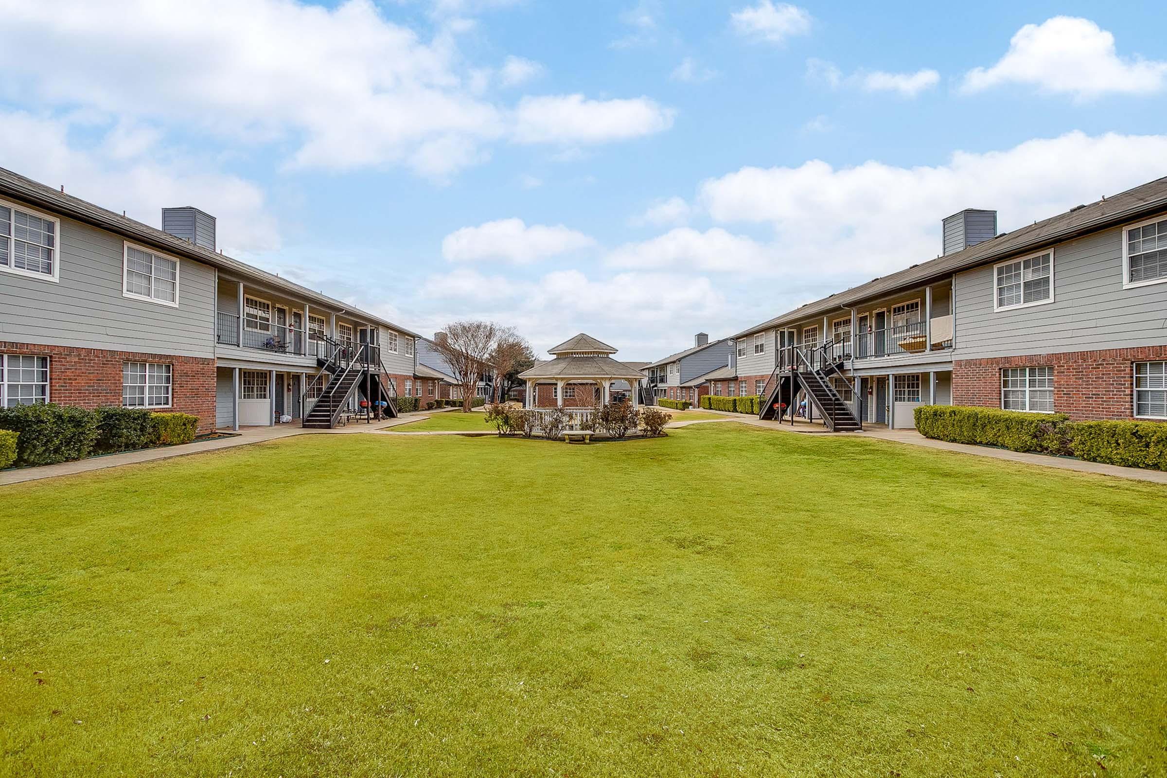 A landscaped courtyard featuring two buildings with staircases on either side. In the center, there is a gazebo surrounded by greenery and well-manicured grass under a cloudy sky. The scene conveys a peaceful residential atmosphere.