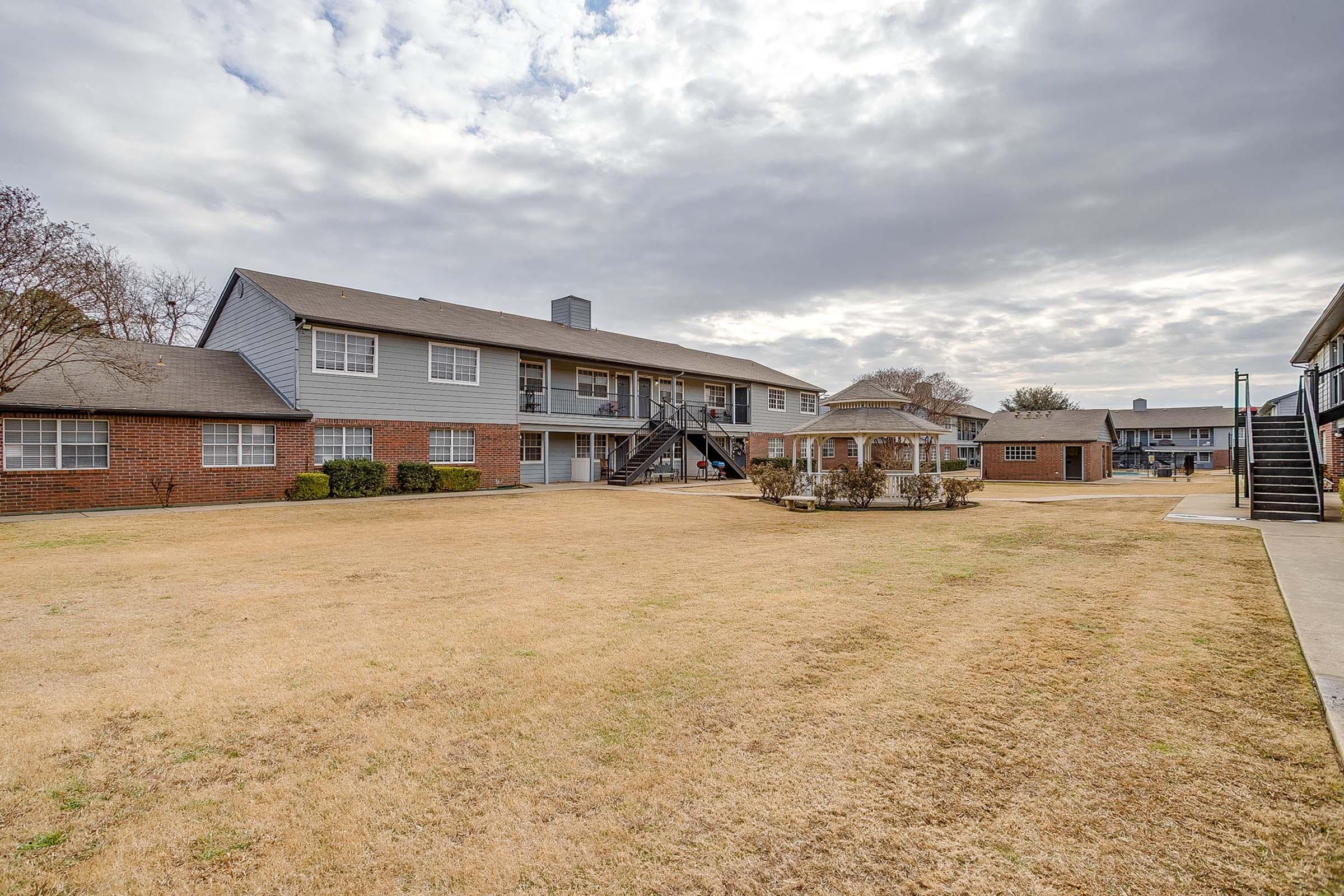 A view of a two-story apartment complex with a grassy courtyard, featuring a gazebo and a staircase leading to the upper level. The sky is partly cloudy, and the surrounding area includes additional buildings and landscaping.