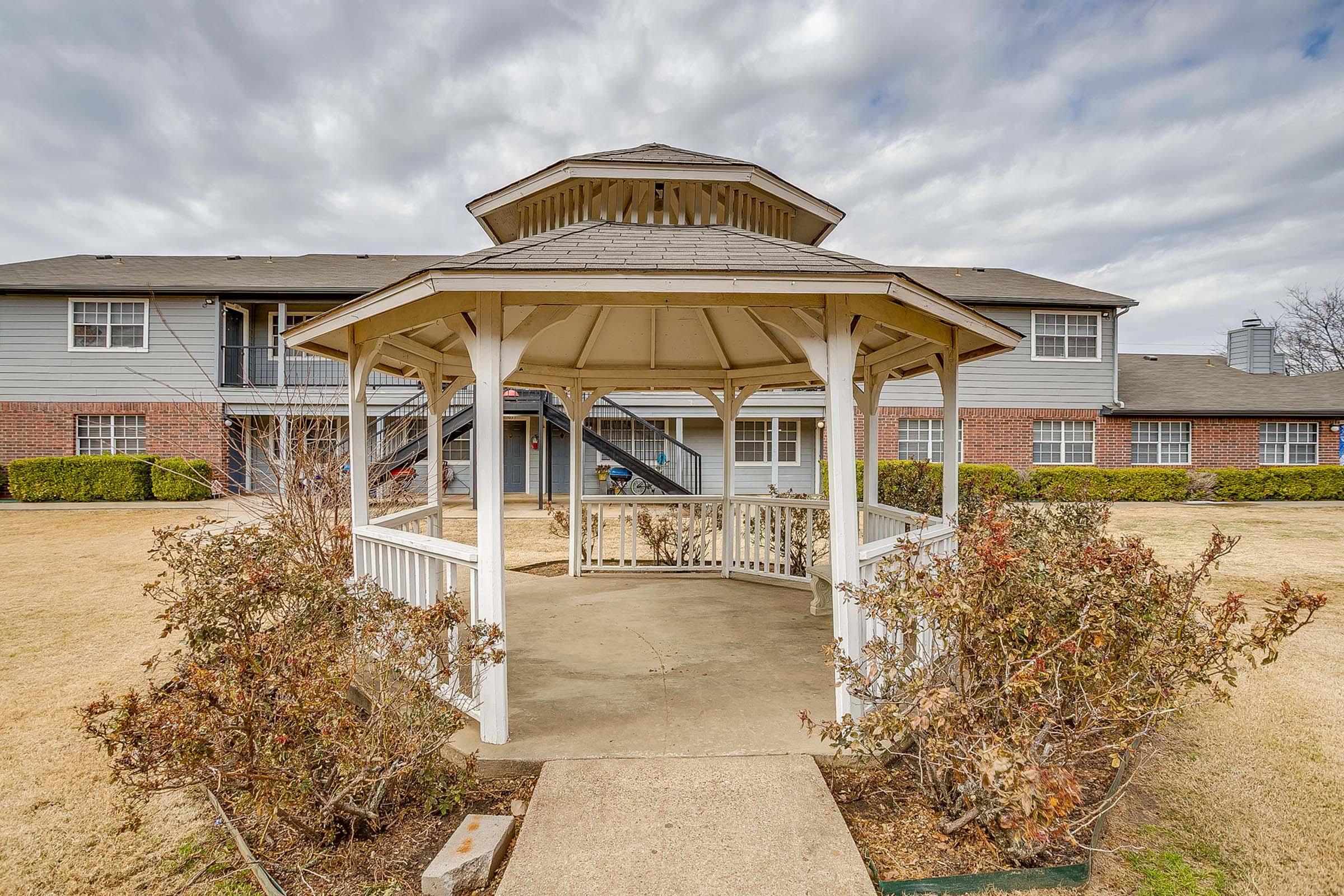 A white wooden gazebo surrounded by barren shrubs, with a pathway leading to it. In the background, there are two-story apartment buildings and a cloudy sky overhead. The setting appears to be a landscaped area in a residential complex. The grass is light brown, indicating a potentially dry season.