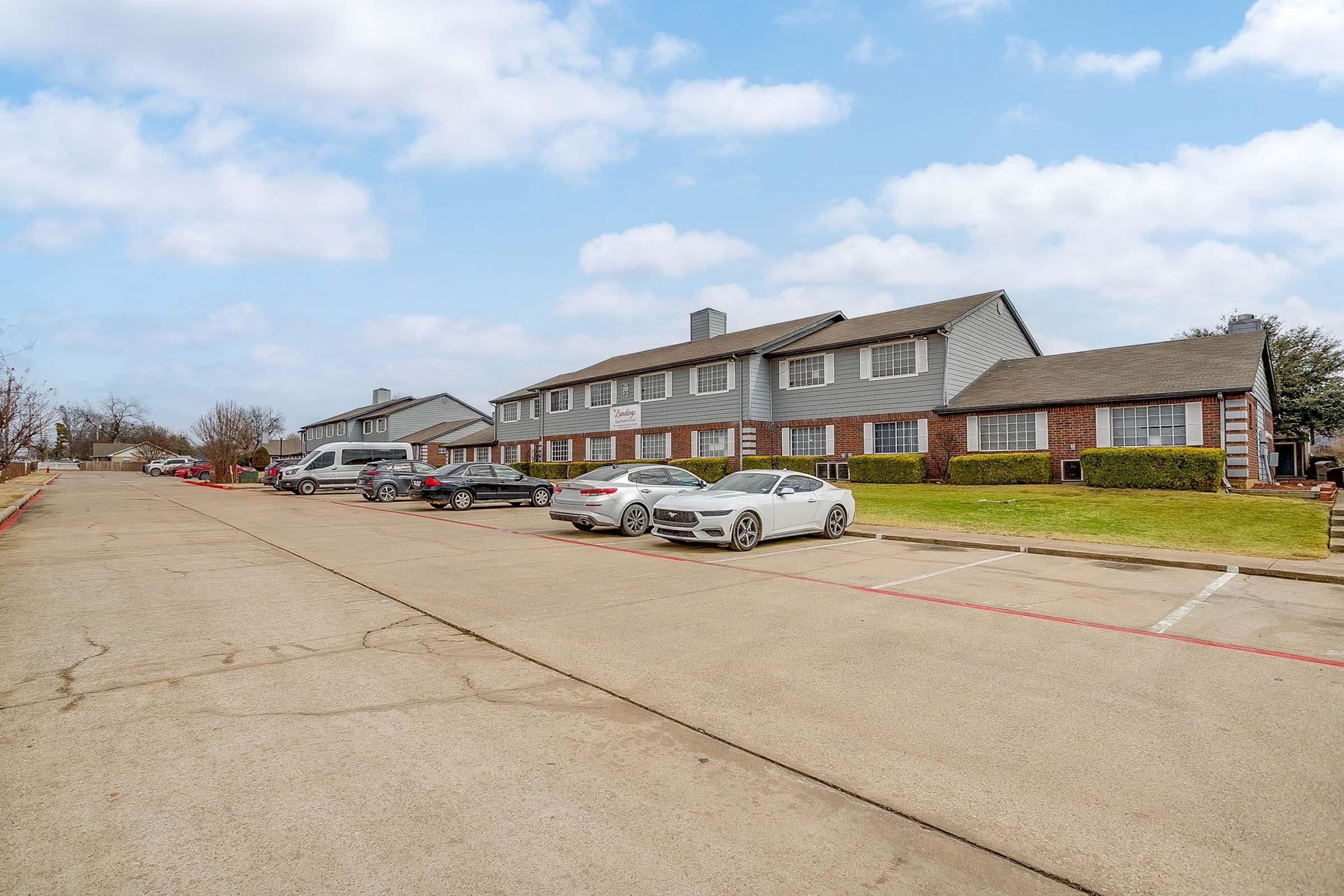 A view of a residential area featuring a row of townhouses with a paved parking lot. Several cars are parked in front of the buildings, and there are patches of grass and trees lining the sidewalk. The sky is partly cloudy, creating a bright, inviting atmosphere.