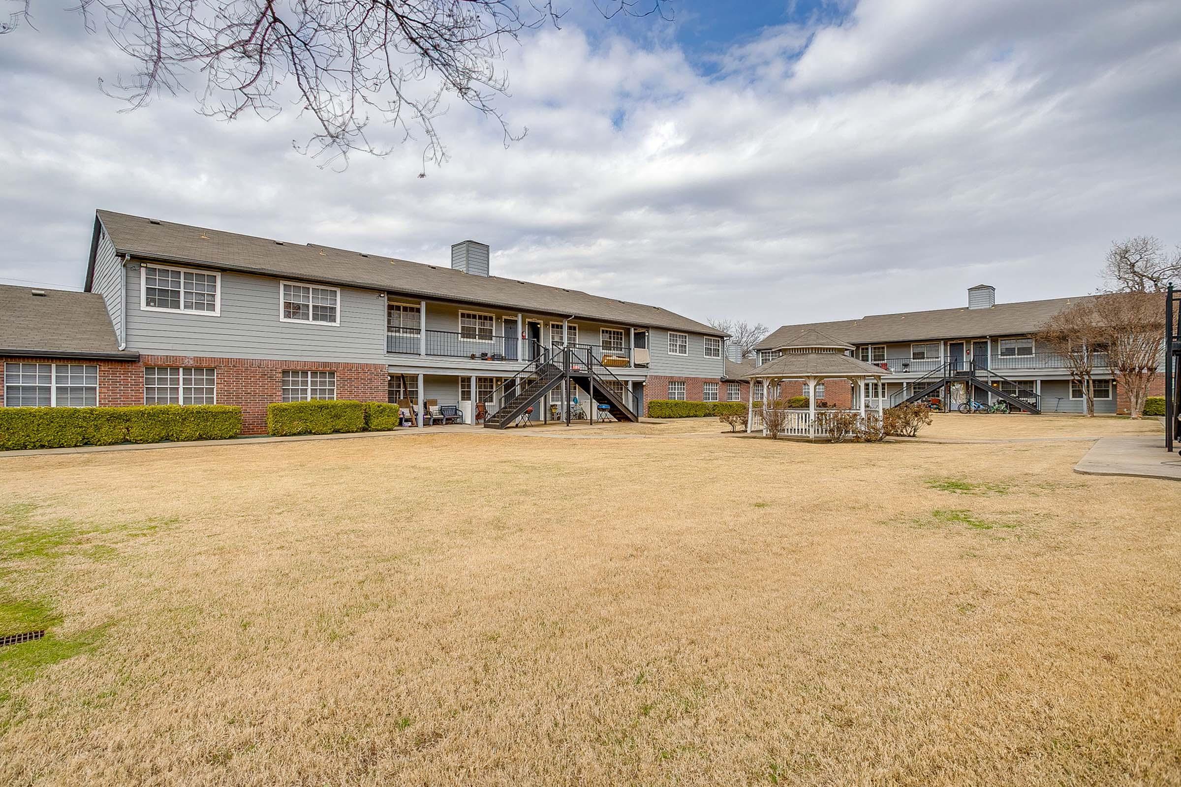 A view of two multilevel apartment buildings with a grassy area in the foreground. The buildings feature balconies and are surrounded by trimmed bushes. The sky appears overcast with clouds.