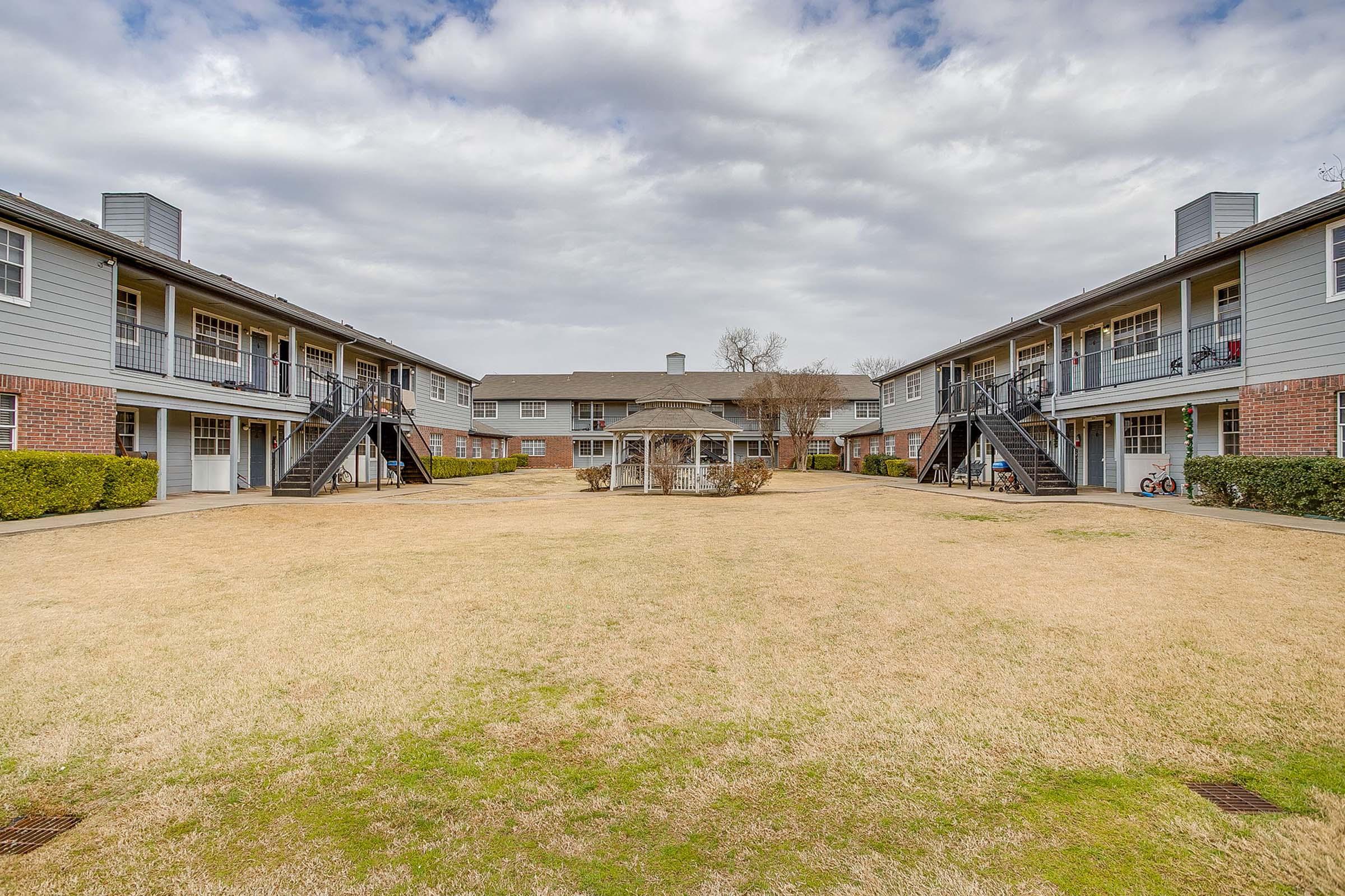A view of a residential apartment complex featuring two-story buildings surrounding a central grassy courtyard. There are wooden staircases leading to the upper units, with a gazebo in the middle and several bicycles parked nearby. The sky is overcast, contributing to a serene atmosphere.