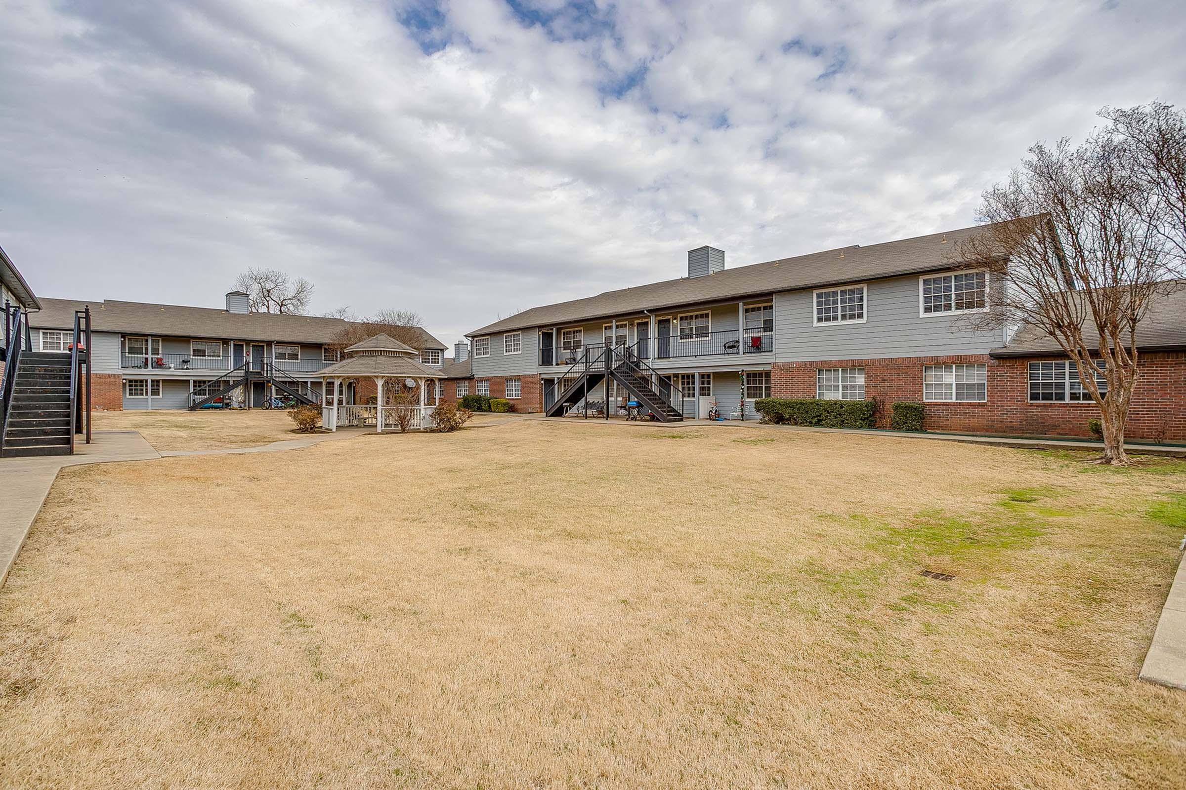 A view of a spacious apartment complex with multiple two-story buildings. The foreground features a dry, grassy area, while the background shows well-maintained pathways and a covered pavilion. The sky is partly cloudy, suggesting an overcast day.