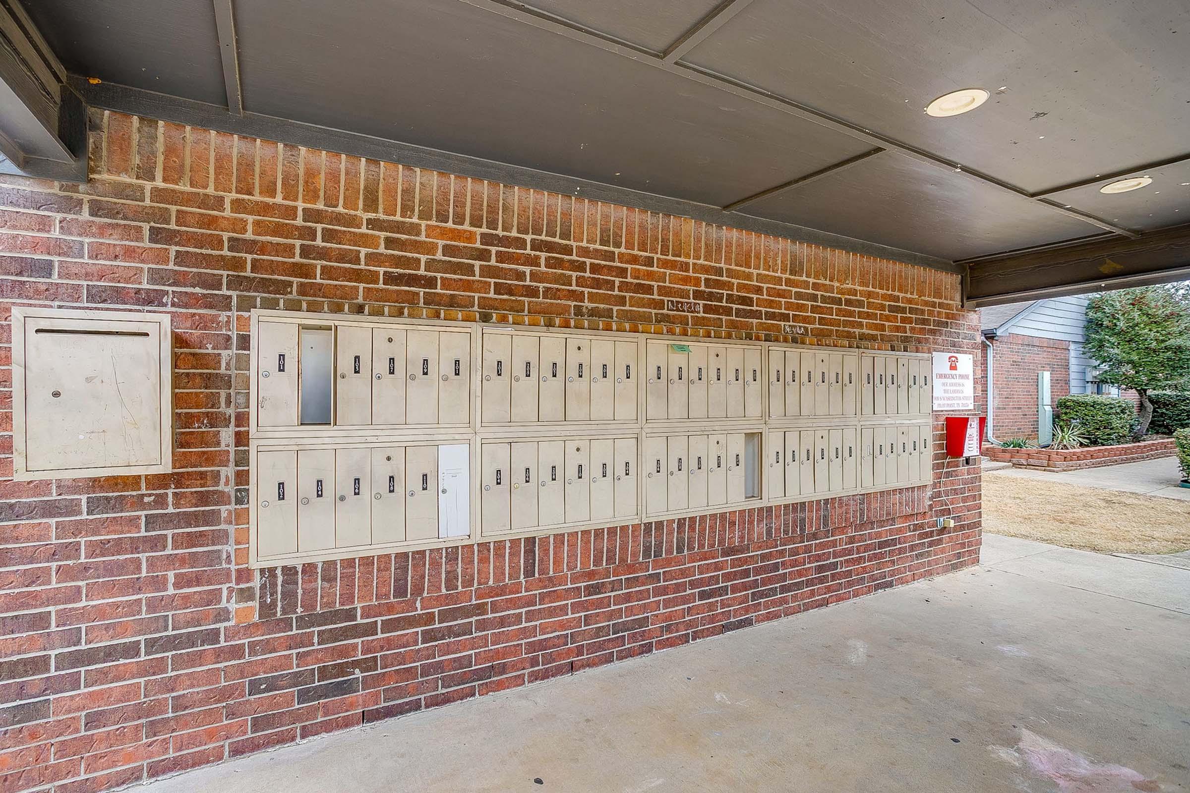 Row of metal mailboxes mounted on a brick wall under a covered area. The mailboxes are neatly arranged, with some displaying numbers. A single mail drop box is positioned to the left. The ground is concrete, and the surrounding area has dry grass and landscaping.