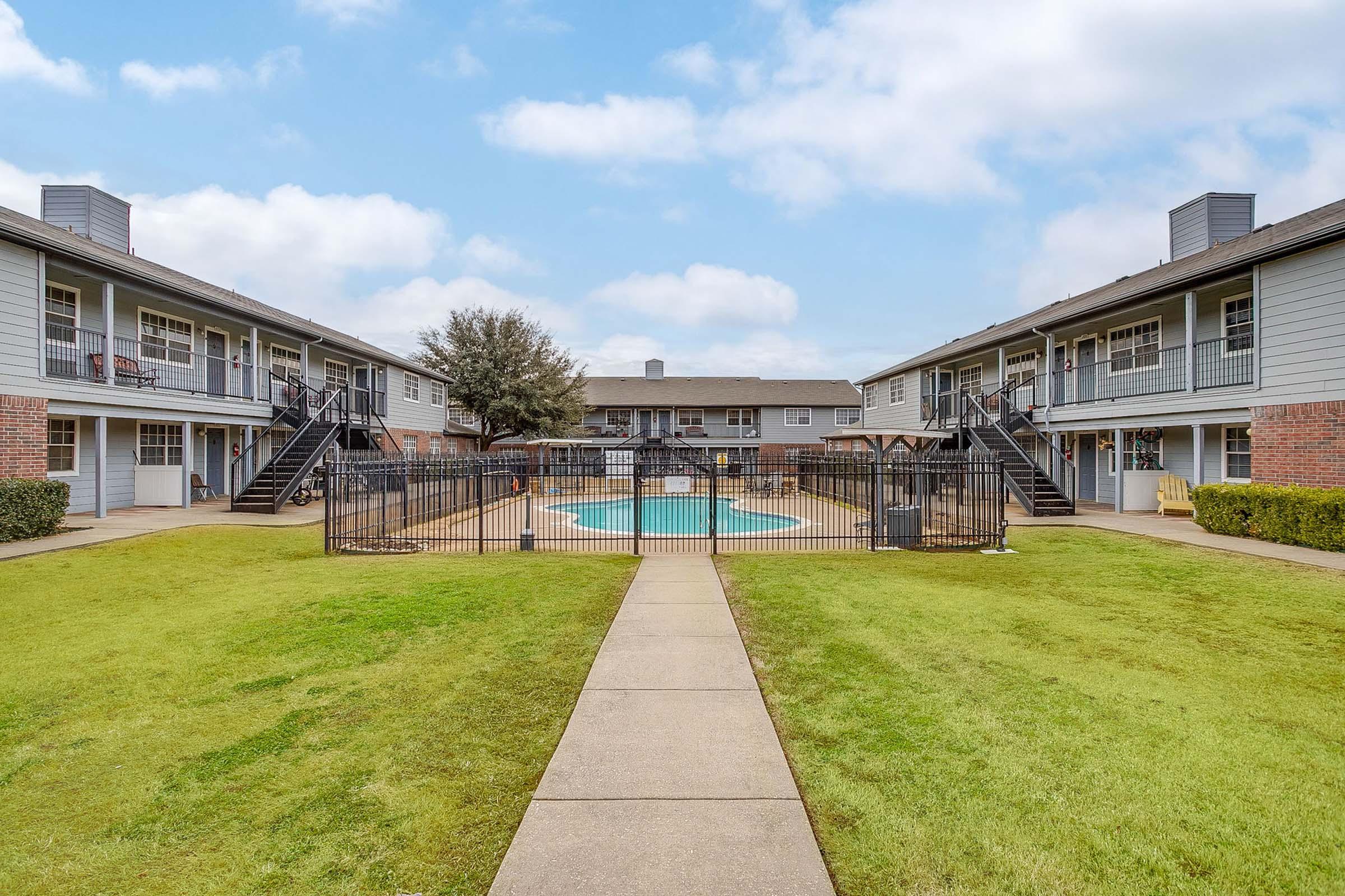 A landscaped courtyard scene featuring two multi-story apartment buildings surrounding a fenced swimming pool. There are pathways leading to the pool area, grassy lawns, and outdoor seating areas visible on balconies. The sky is partly cloudy, creating a pleasant atmosphere.
