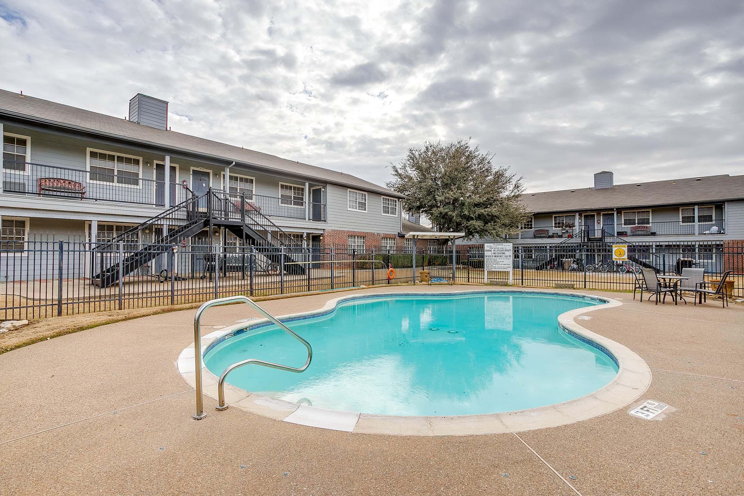 A swimming pool surrounded by a fenced area, with lounging chairs nearby. Two-story apartment buildings are in the background under a cloudy sky. A staircase leads to the apartments, and there are signs by the pool area. The scene conveys a peaceful residential atmosphere.