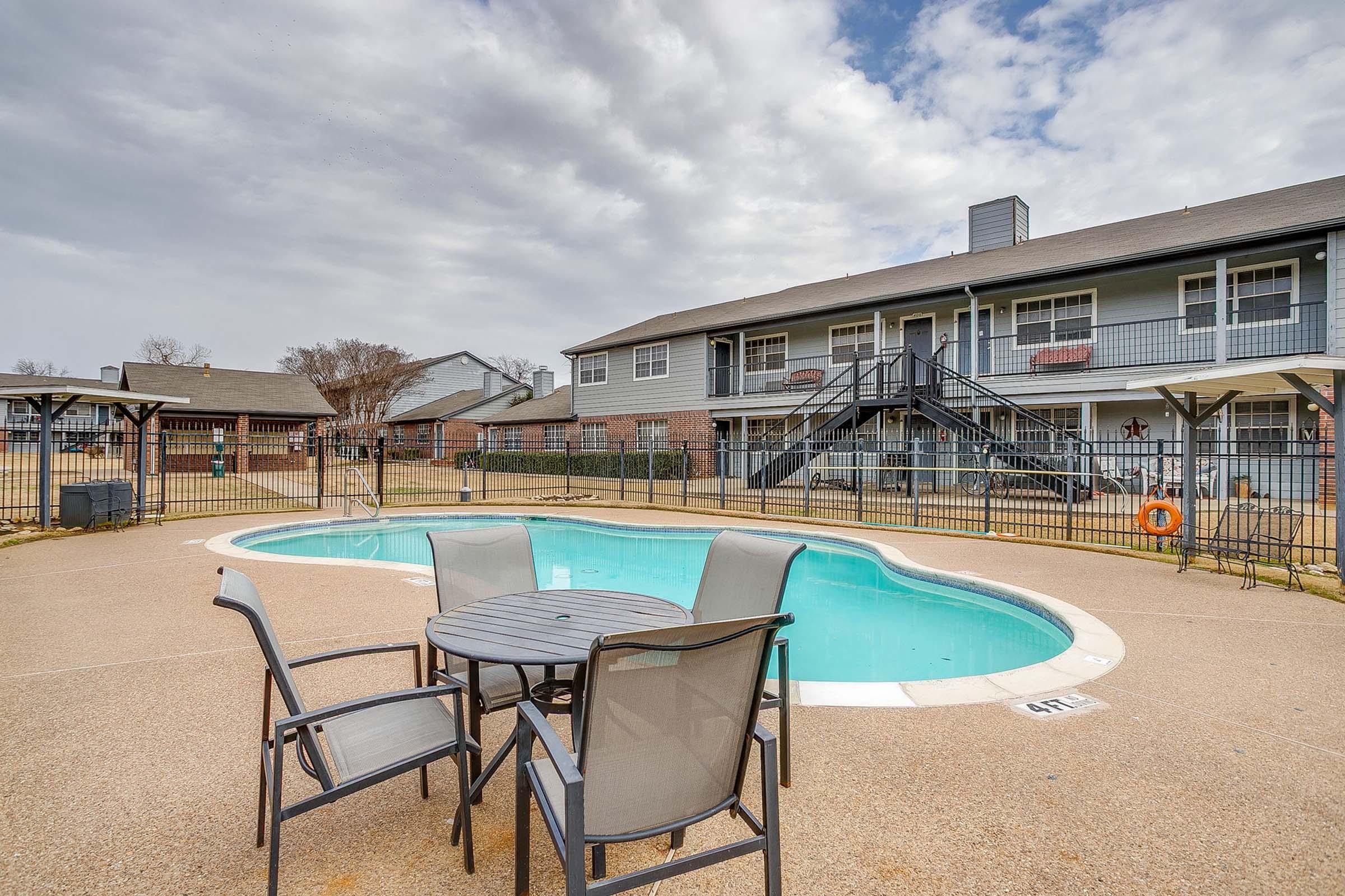 A view of a swimming pool surrounded by a concrete area featuring a small table and chairs. In the background, there are two-story apartment buildings with balconies, and the sky is partly cloudy. The pool area is fenced, and there are shaded seating areas visible.