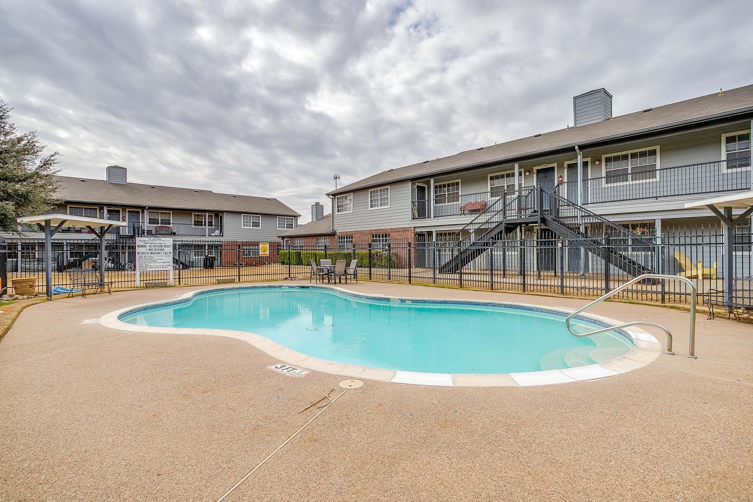 A well-maintained outdoor swimming pool surrounded by a fence, located in an apartment complex. The pool area features a diving board and lounge chairs, with two multi-story buildings in the background and a cloudy sky overhead.